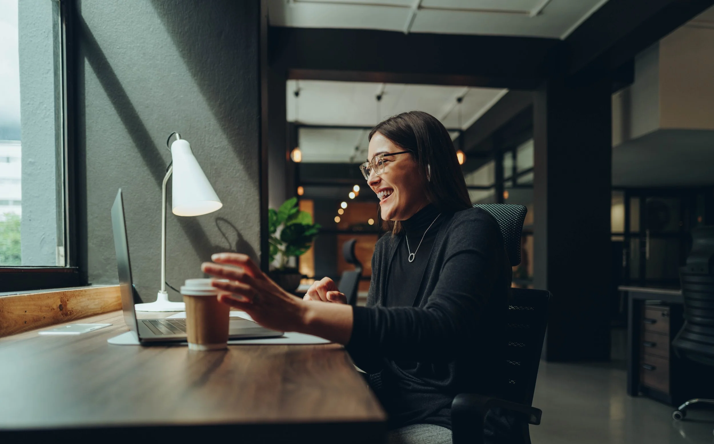 A woman with glasses and dark hair sitting at a desk in a modern office, smiling while looking at her phone with a coffee cup in front of her, near a laptop, and a lamp on the desk, with large windows and greenery outside.