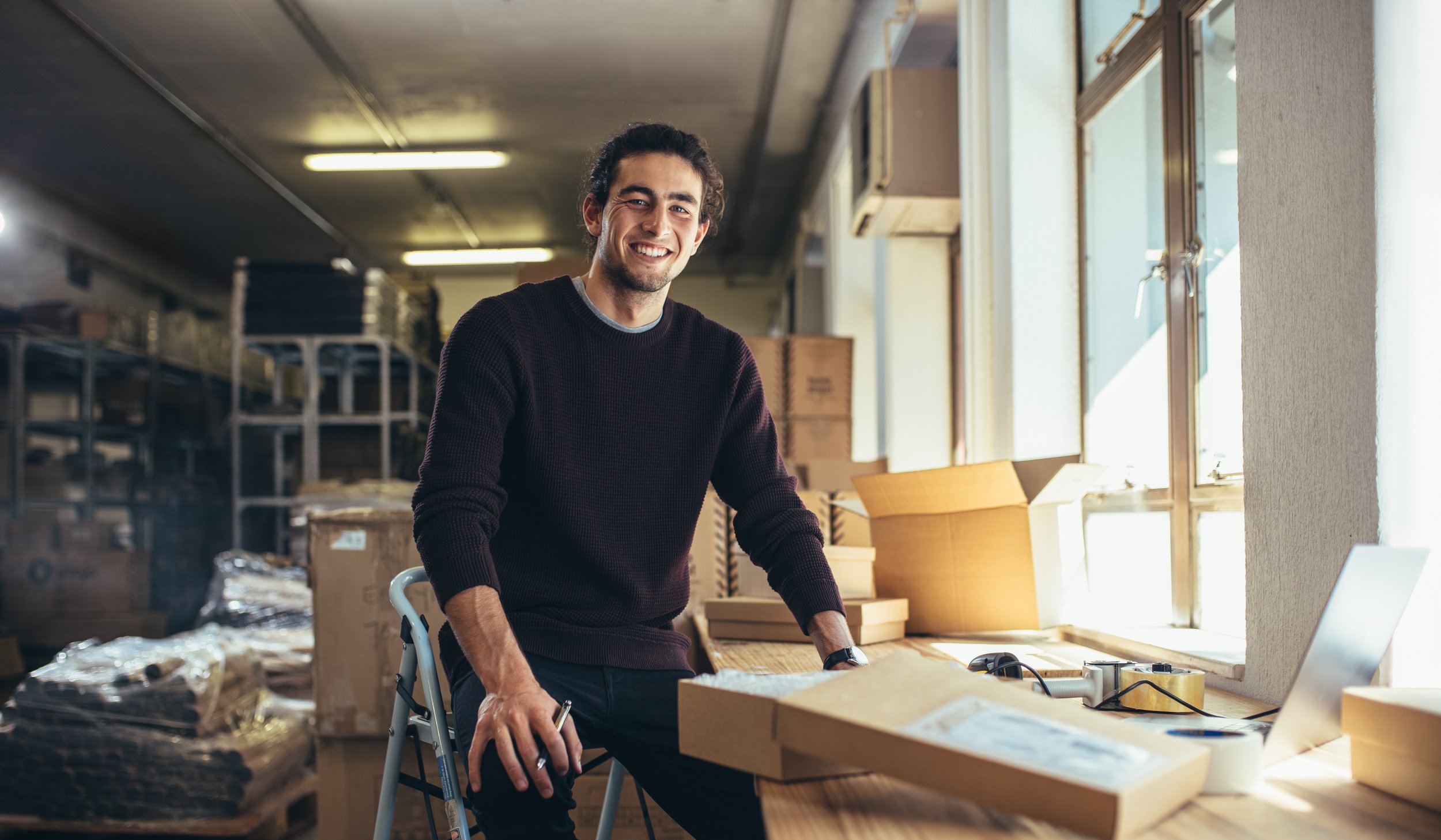 Smiling young man in a dark sweater sitting at a workbench in a workshop surrounded by cardboard boxes and tools, looking at the camera.