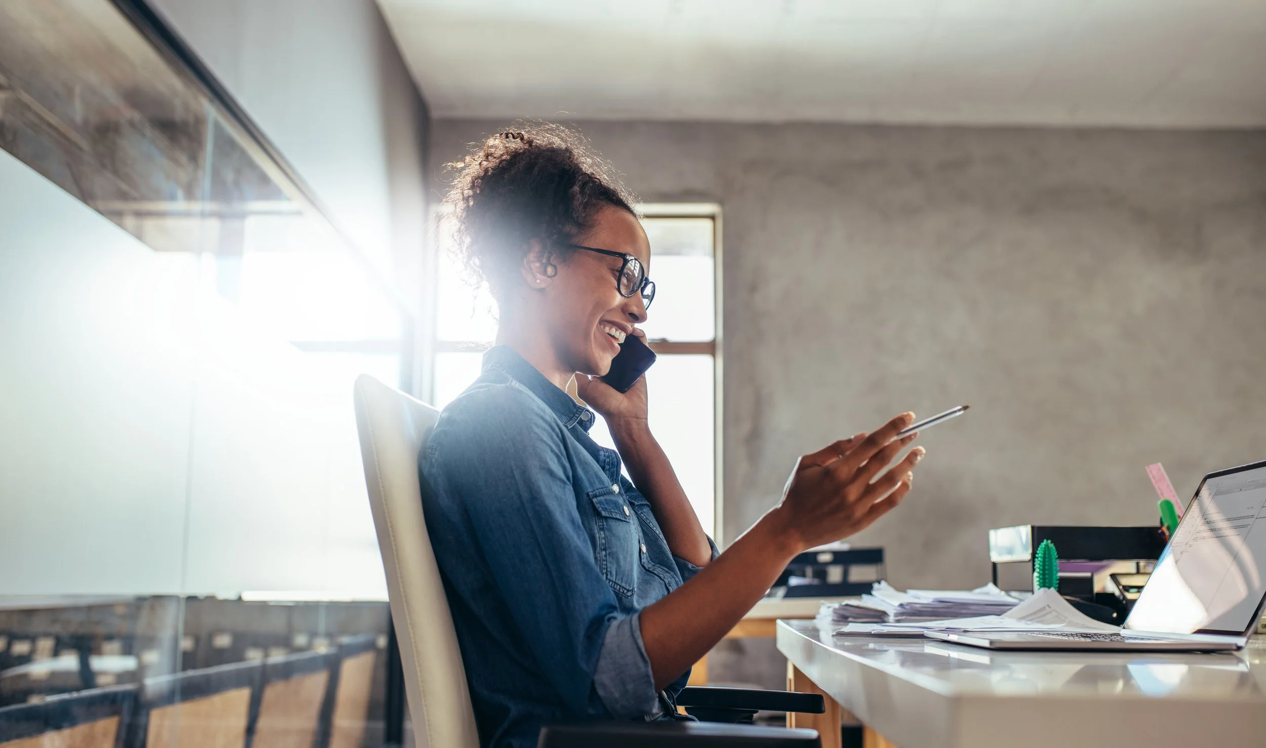A woman with curly hair, glasses, and a denim shirt smiling while talking on her cellphone in a modern office with natural light, desk clutter, and a laptop.