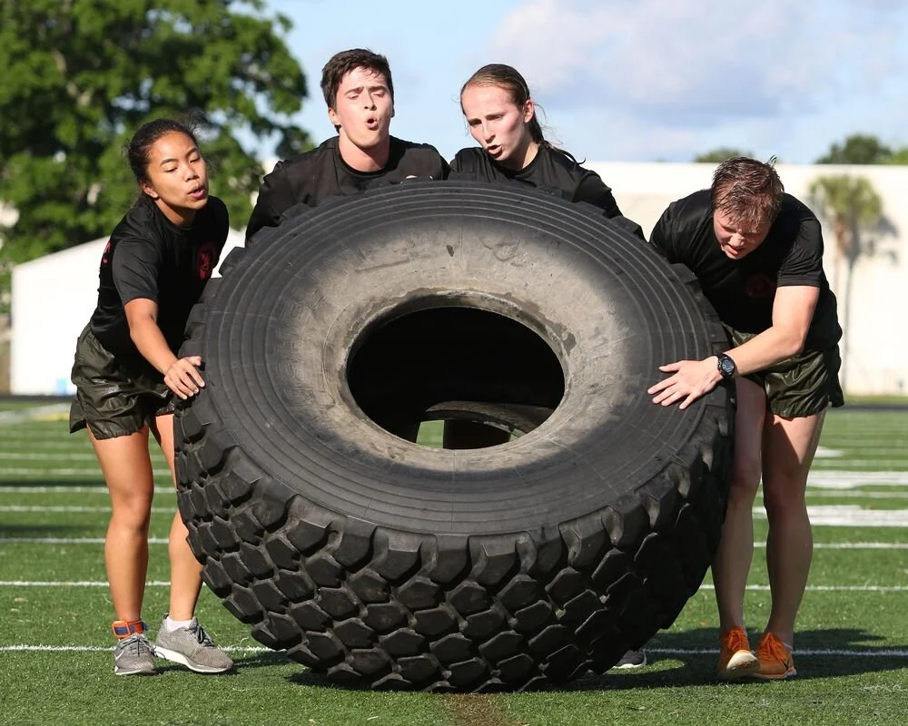 Four young athletes in black uniforms perform a tire flip exercise outdoors on a grassy field with white lines, trees, and a cloudy sky in the background.
