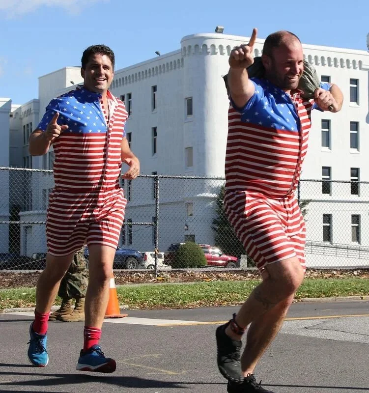 Two men running in a race, wearing matching American flag-themed outfits, one giving a thumbs-up and the other raising a finger in the air, with a white building in the background.
