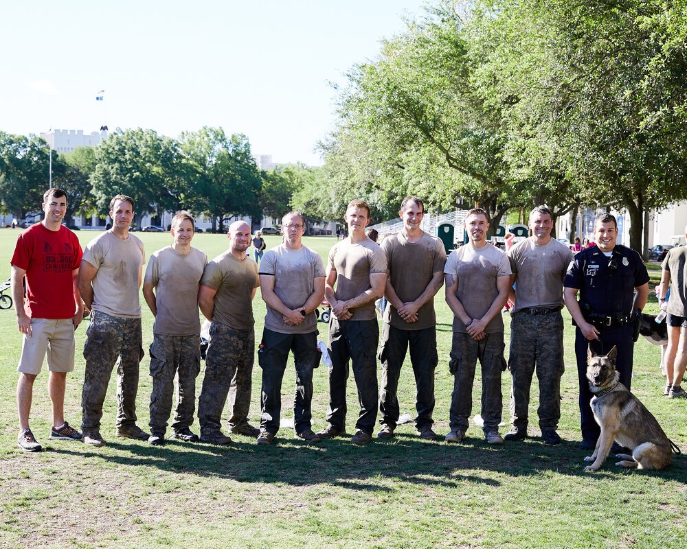 Group of nine men, mostly in military camouflage pants and shirts, standing on grass with a police officer and a German shepherd dog. Trees and a building are in the background on a sunny day.