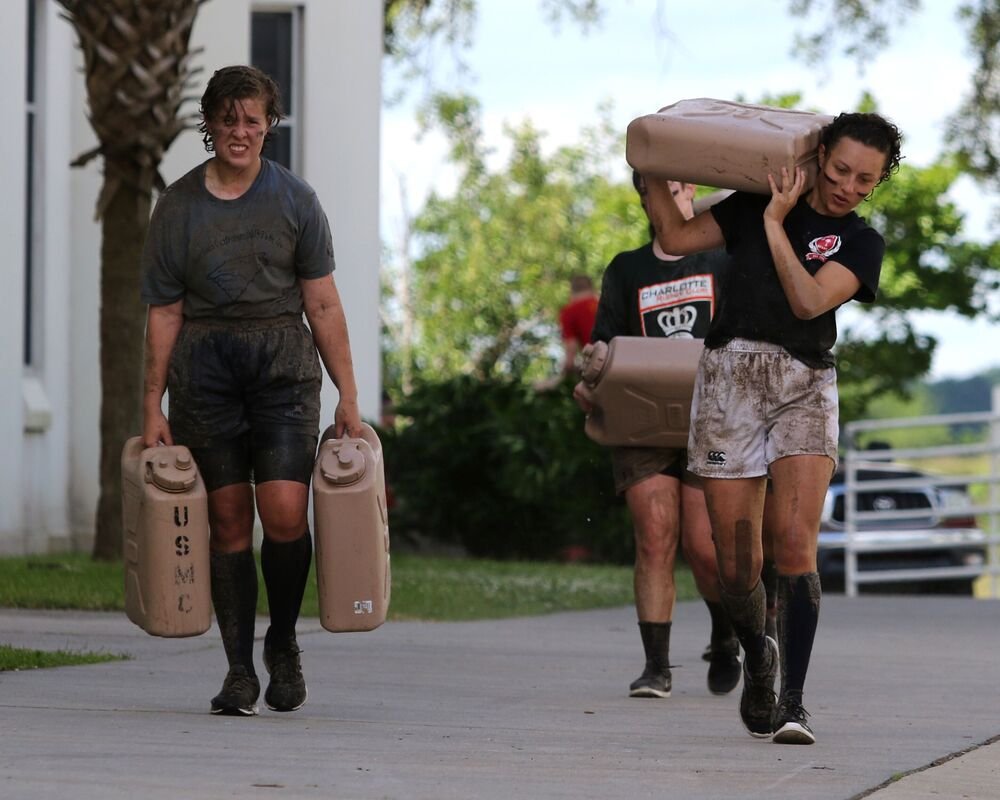 Three women covered in mud carrying jerry cans outside.
