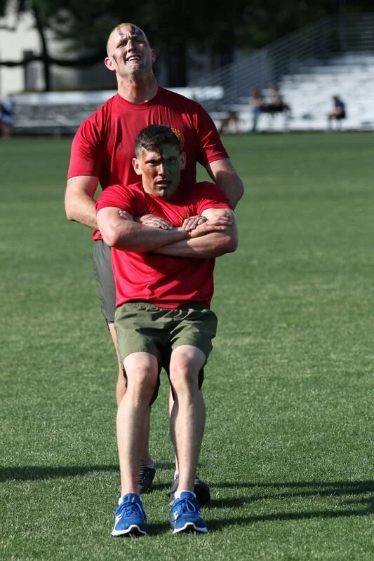 Two men in red shirts and shorts practicing strength training on a grassy field, with stadium seating and trees in the background.