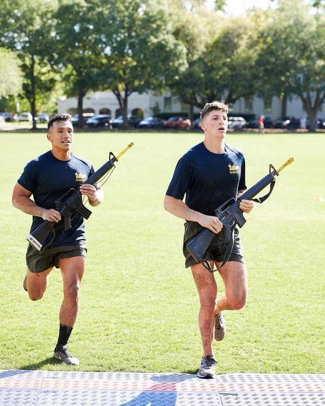 Two young men running outdoors on a grassy field while carrying toy guns, with trees and parked cars in the background.