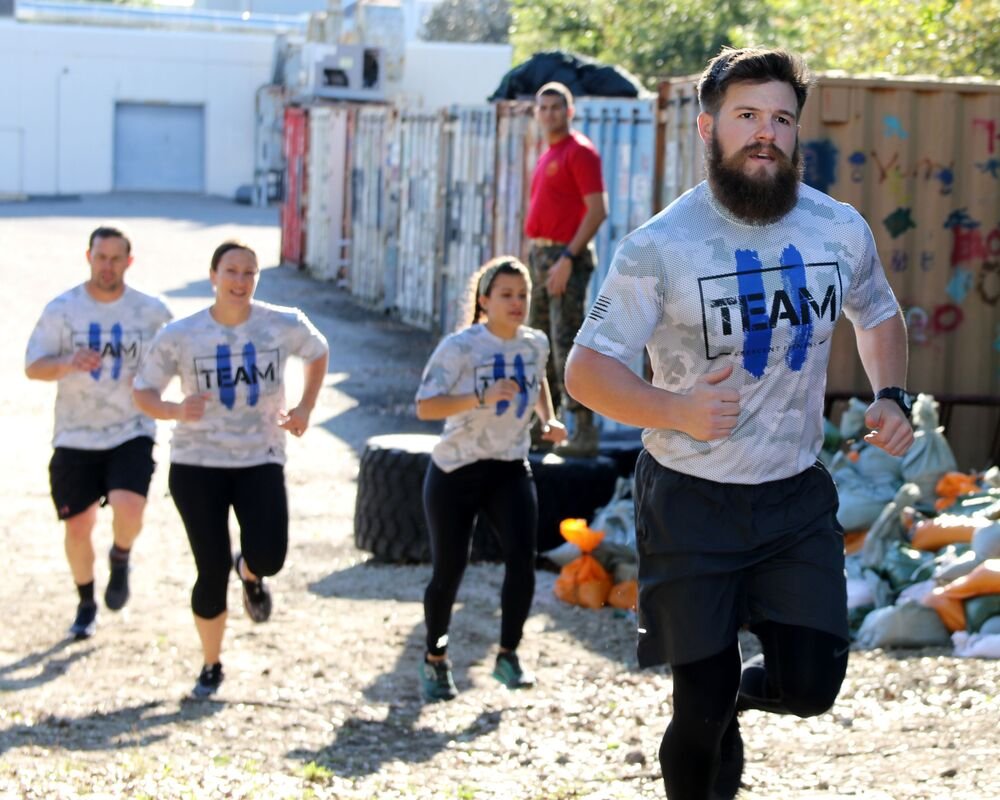 A group of four runners, wearing matching white and gray camouflage shirts with "TEAM" printed on them, running outdoors on a sunny day, with a man in a red shirt standing in the background near a fence.