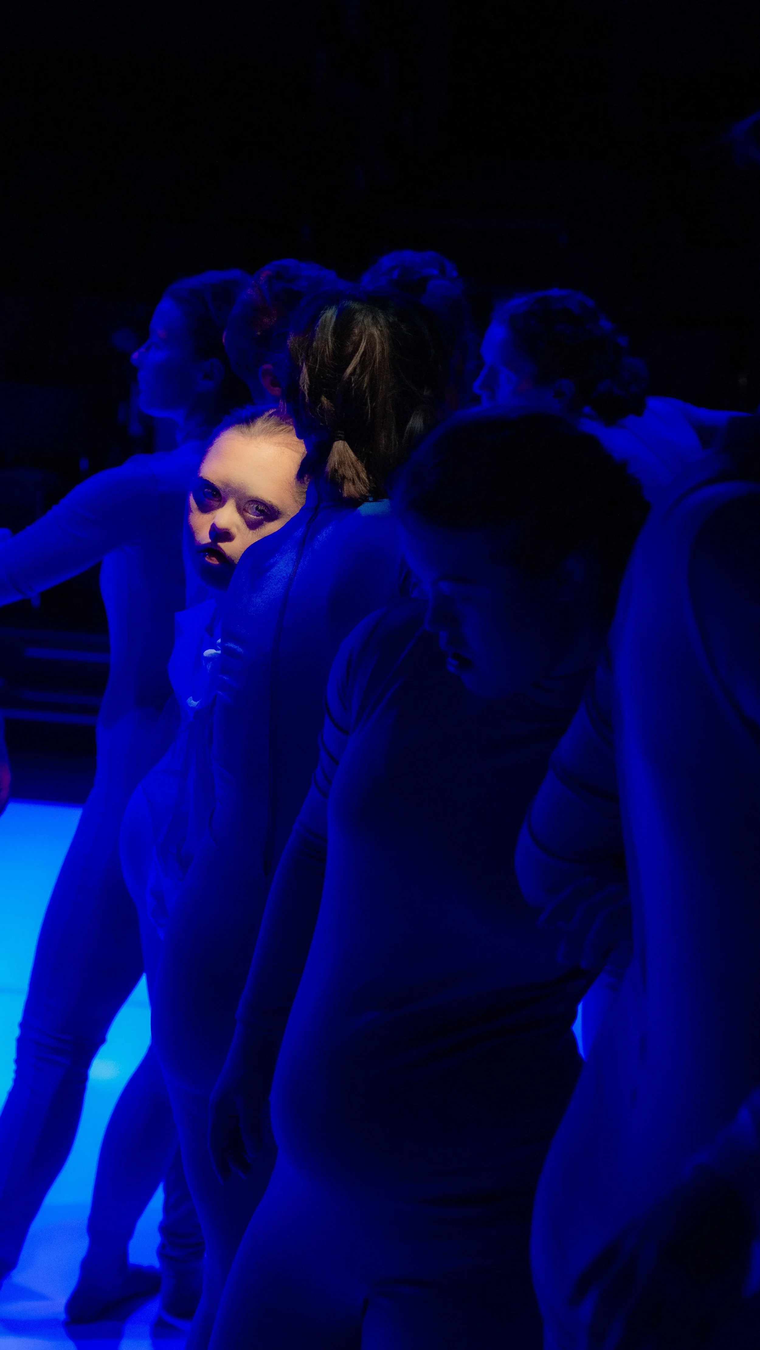 Menschen in dunklem Raum, hauptsächlich in Blau beleuchtet, stehen in einer Reihe, eine Person blickt direkt in die Kamera.