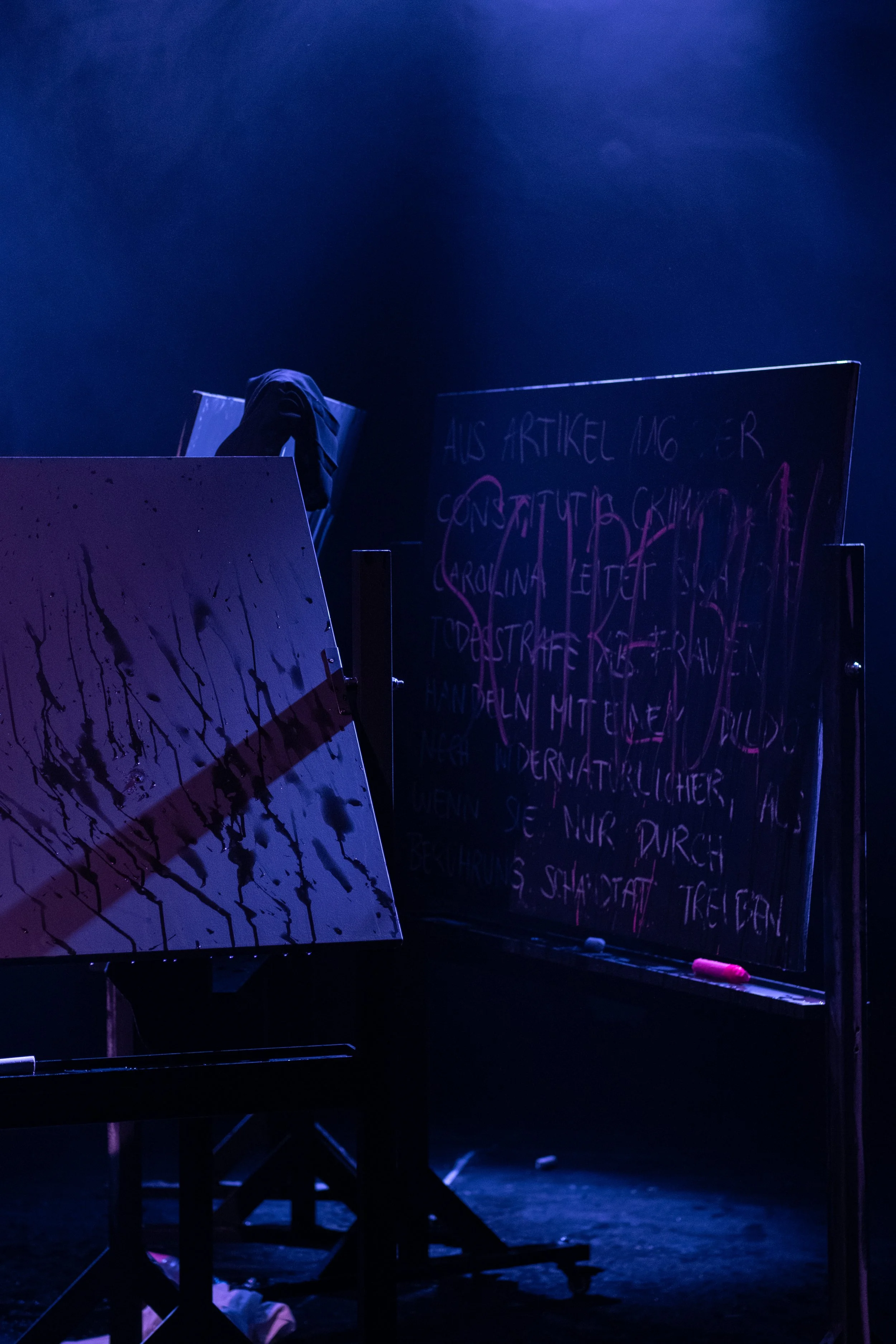 Ein dunkler Bühnenbereich mit einem schwarzen Tafel, an dem pink schreibende Kreide befestigt ist, und einem Flipchart mit bunter Schrift, der von Scheinwerfern beleuchtet wird.