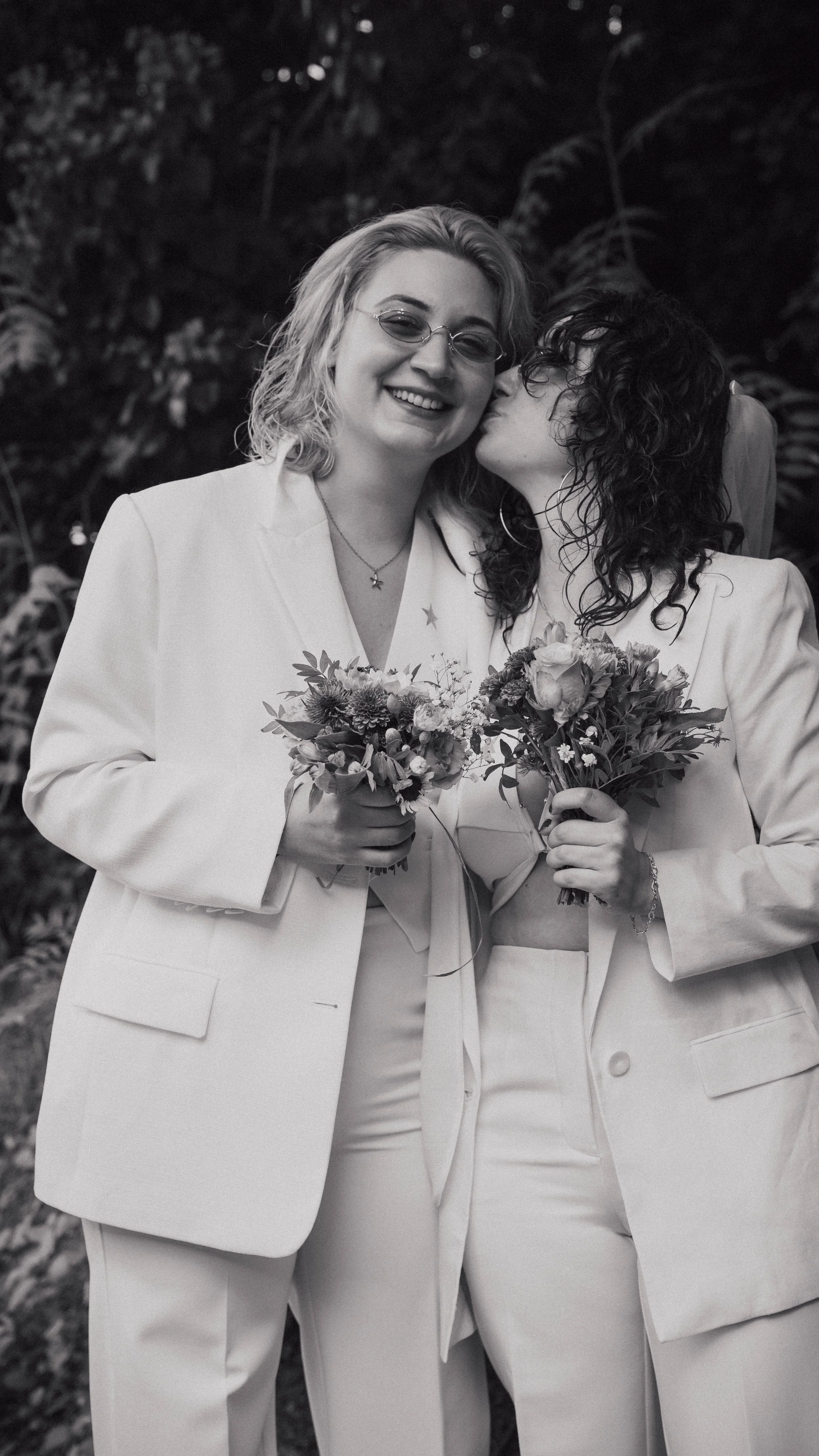 Zwei Frauen in weißen Anzügen, die sich umarmen und Blumen halten, eine küsst die andere auf die Wange, alles in Schwarz-Weiß.