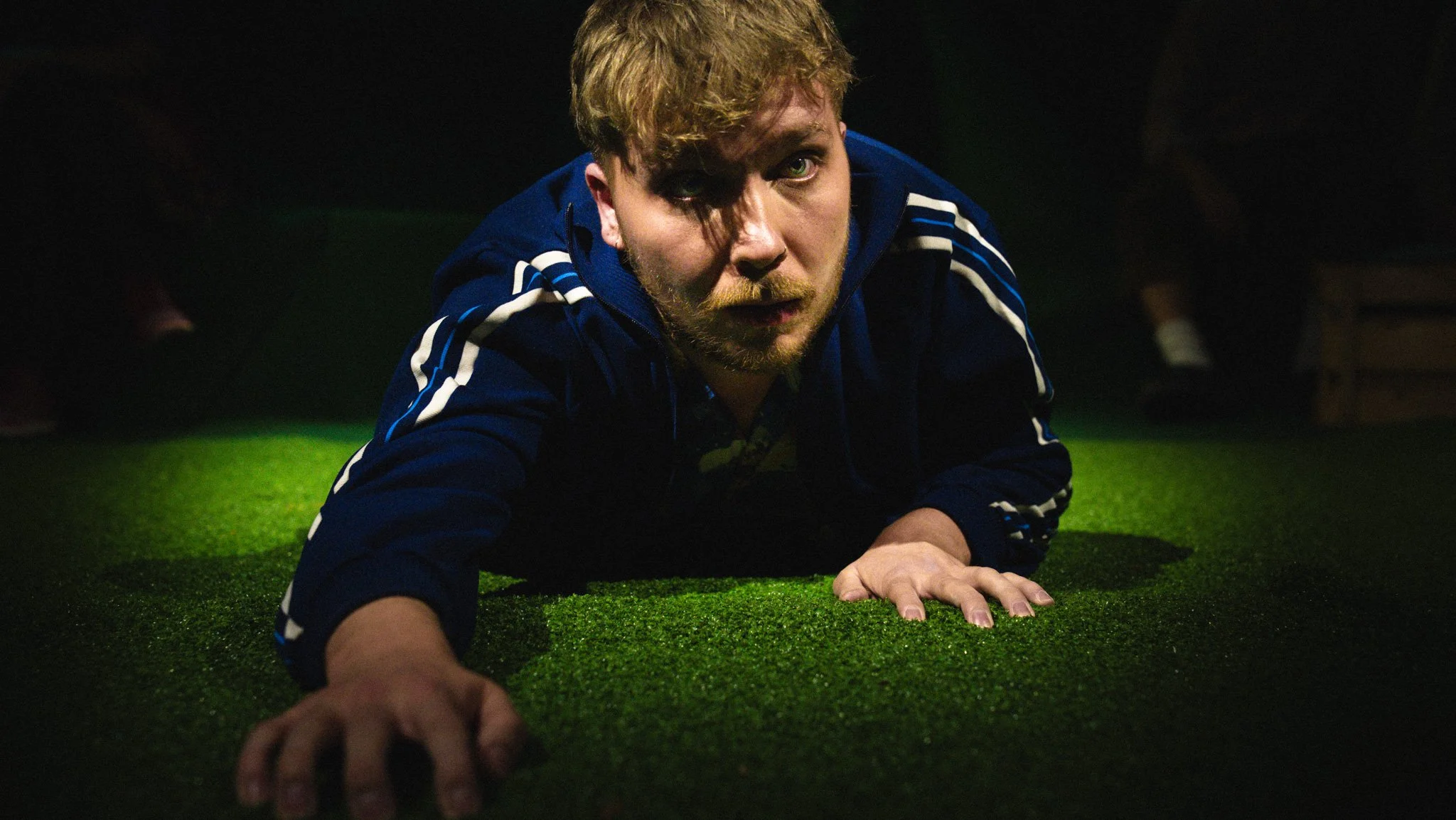 Mann in blauer Jacke auf grünem Rasen liegend in einem Dunkelraum, schaut direkt in die Kamera.