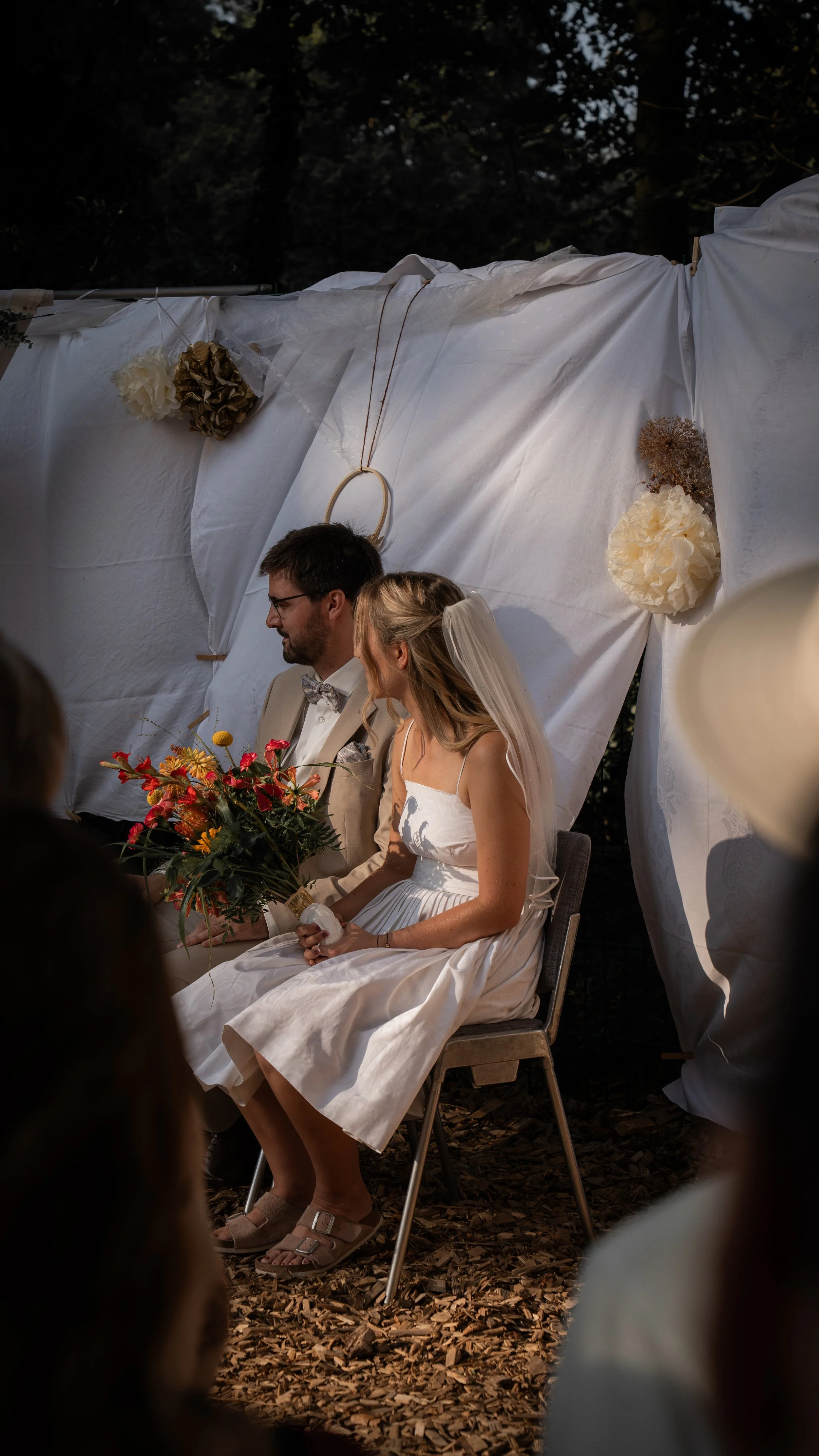 Ein Brautpaar bei einer Hochzeitszeremonie im Freien, sitzend vor einem weißen Dekorationshintergrund mit Blumen und Dekorationen, während sie sich während der Trauung ansehen.