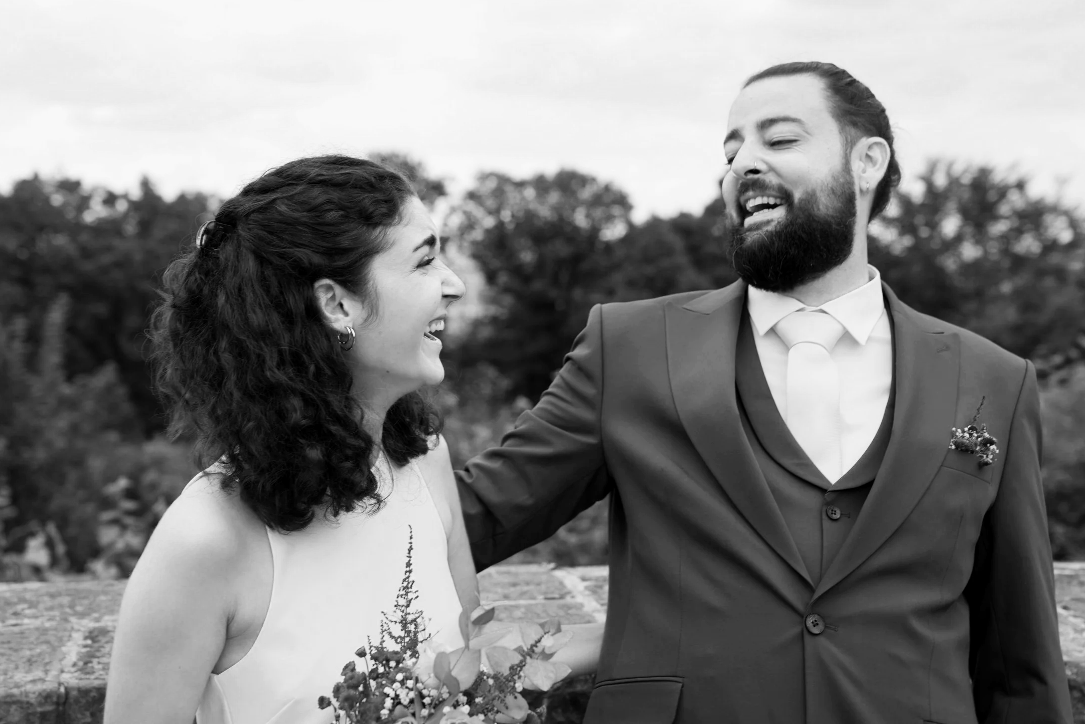 Ein lachendes Paar bei einer Hochzeit, der Mann trägt einen Anzug, die Frau ein Kleid, im Hintergrund Bäume.