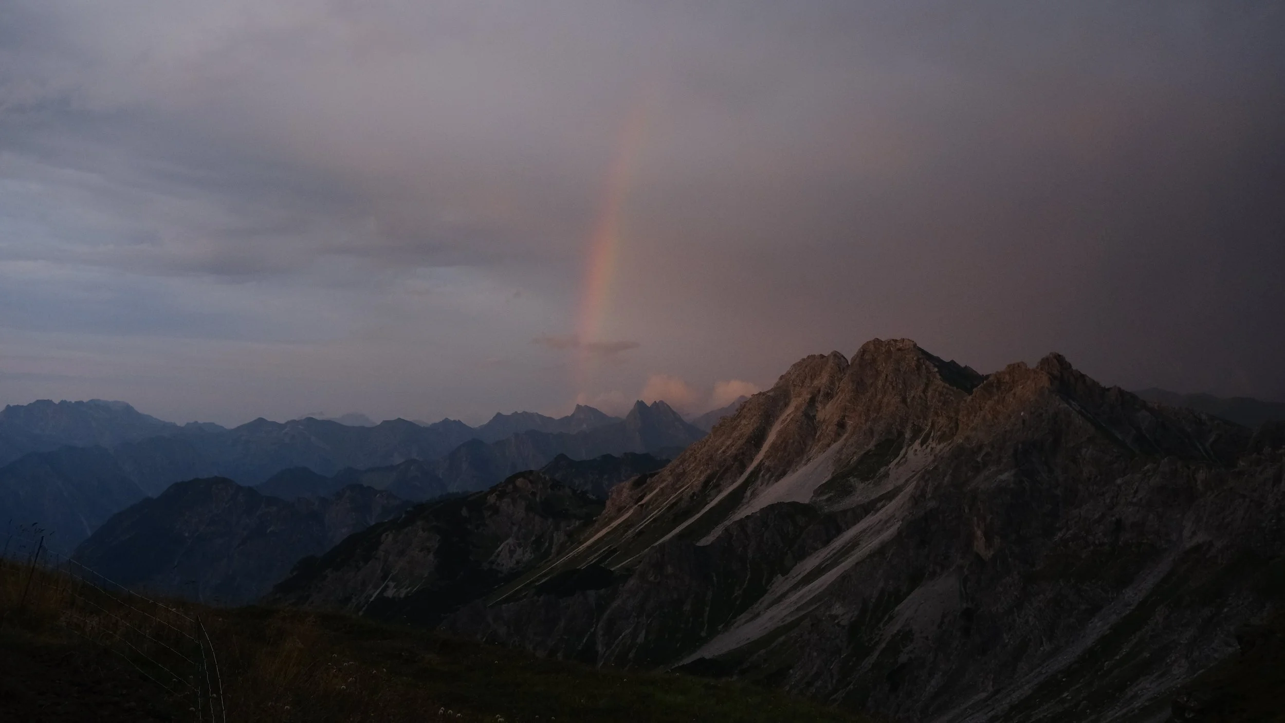 Berge bei Sonnenuntergang mit dunklem Himmel, auf der rechten Seite eine Regenbogen im Himmel.