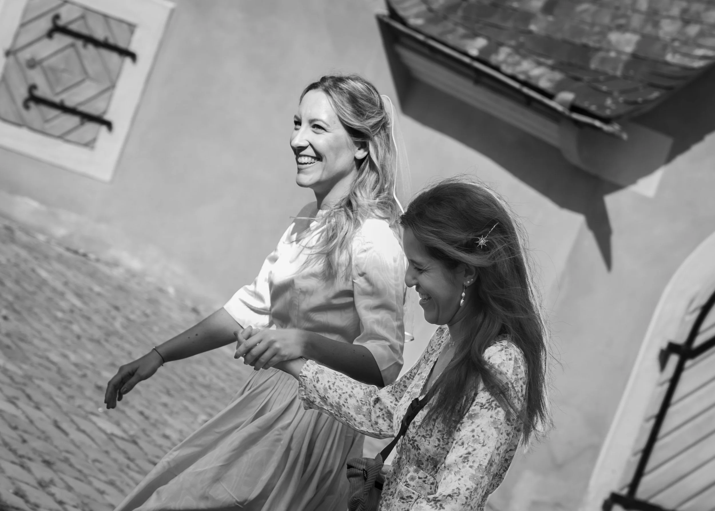 Zwei lachende Frauen, die Hand in Hand laufen, auf einem sonnigen Weg neben einer weißen Wand mit zwei großen Fenstern, eines geöffnet und eines geschlossen.