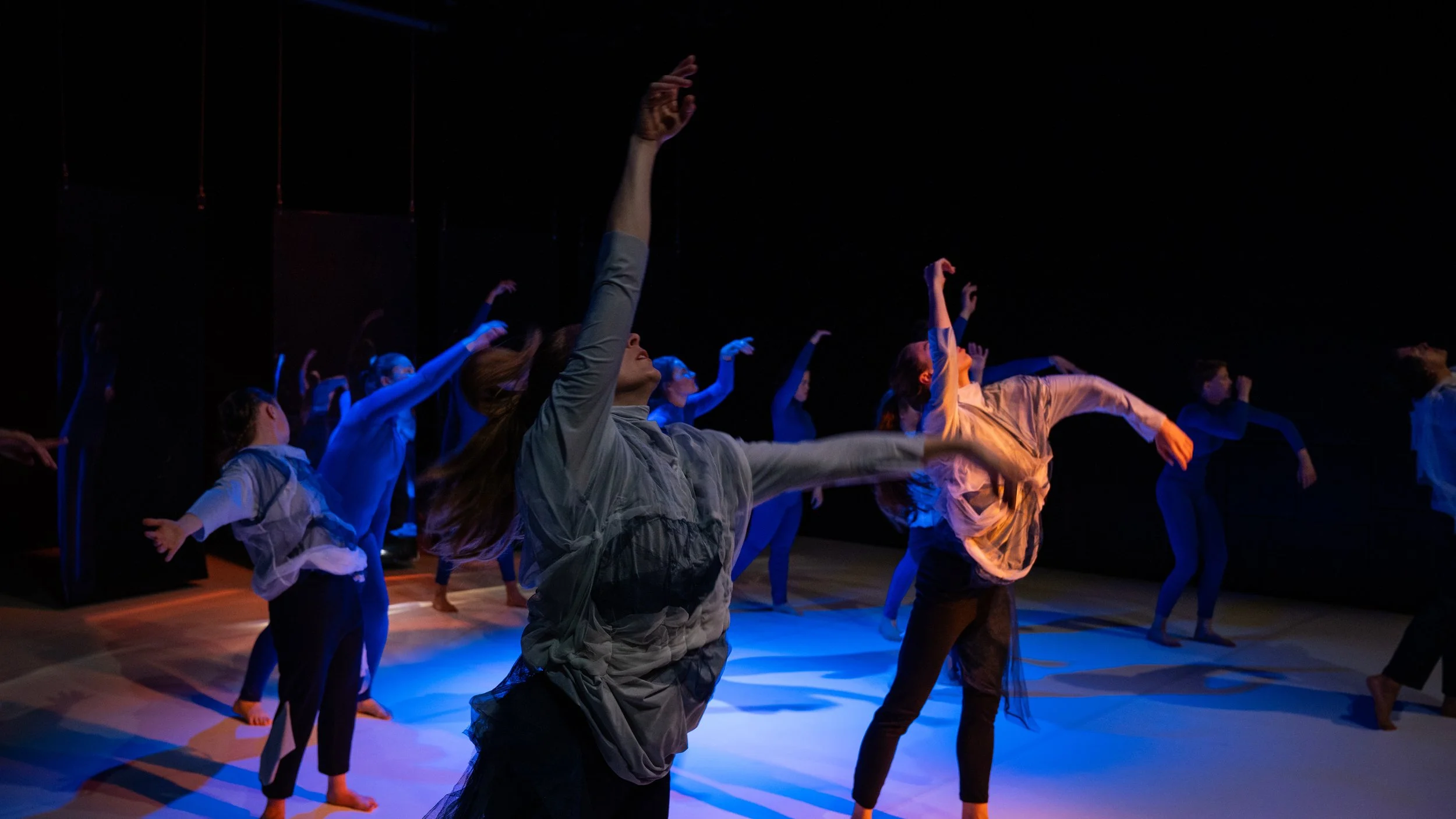 Tanzgruppe auf Bühne, bewegt sich im minimal beleuchteten Raum, mit Fokus auf Bewegung und Ausdruck.