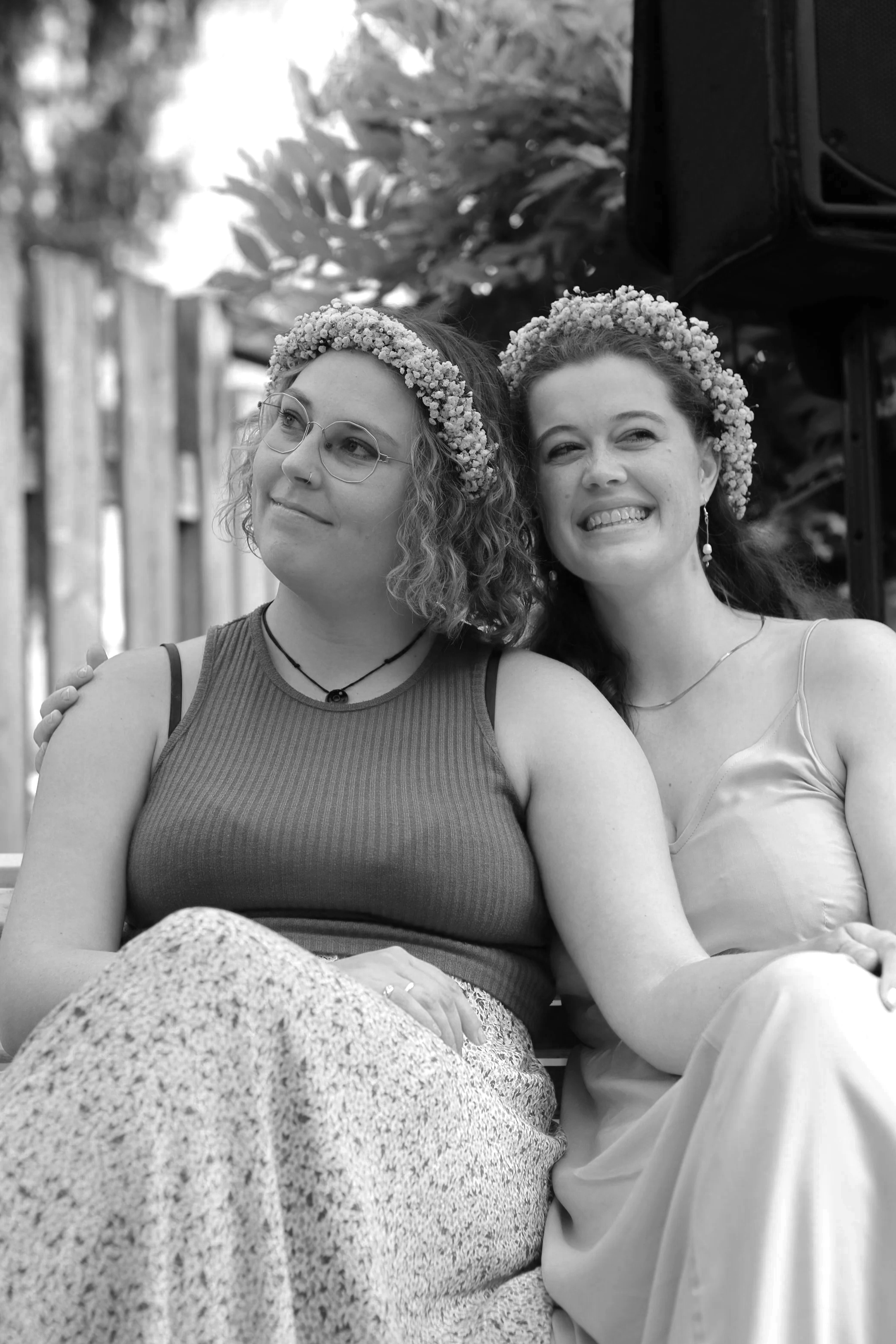 Zwei Frauen sitzen nah beieinander, tragen Blumenkränze im Haar, lachend und lächelnd. Es ist eine schwarz-weiße Aufnahme.