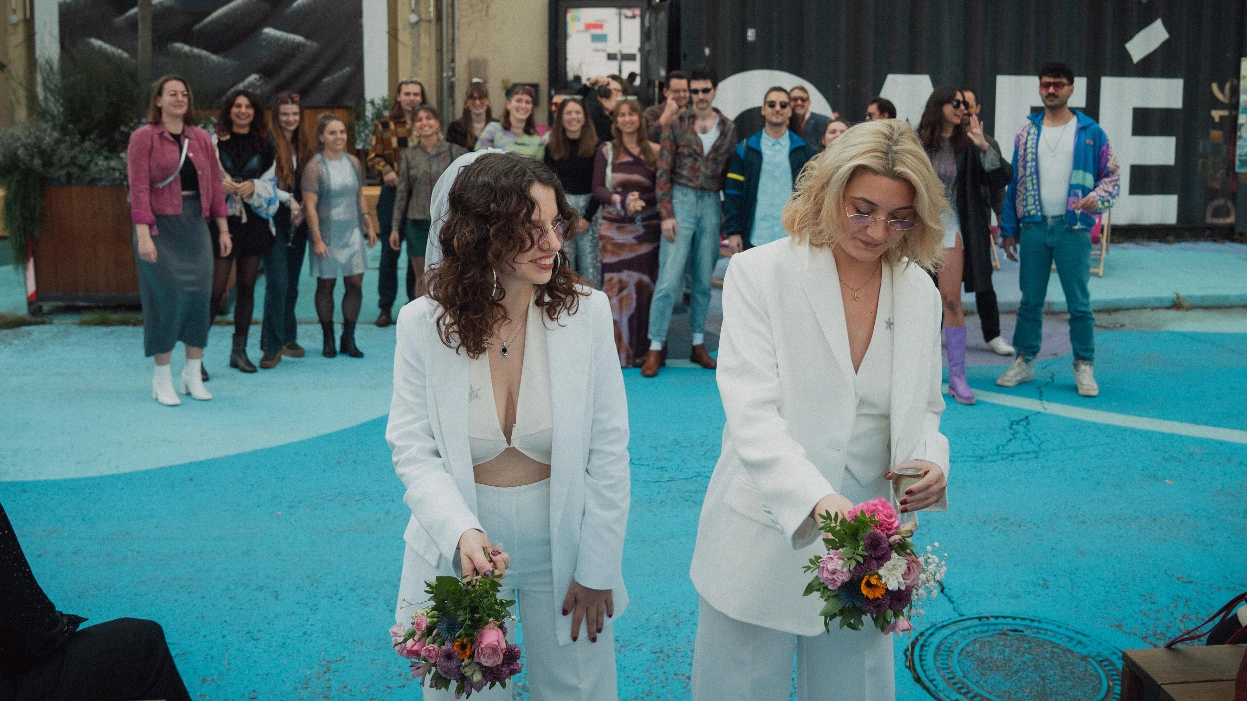 Zwei Frauen in weißen Anzügen mit Blumensträußen vor einer Gruppe von Menschen, die versuchen den Strauß zu fangen.