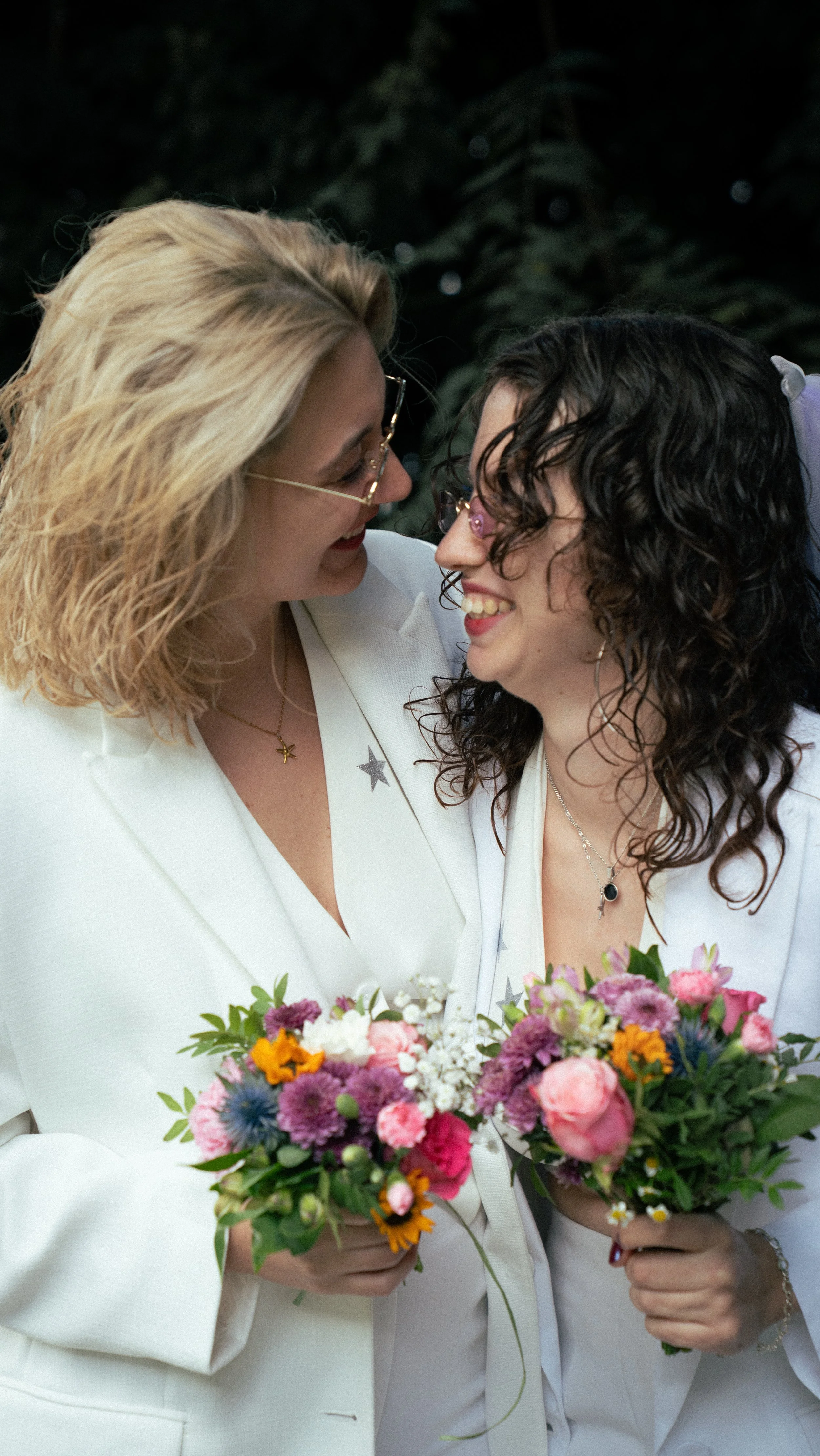 Zwei Frauen in weißen Anzügen, die sich anlächeln und sich mit Blumensträußen umarmen, bei einer Hochzeit oder Feier, im dunklen Hintergrund.