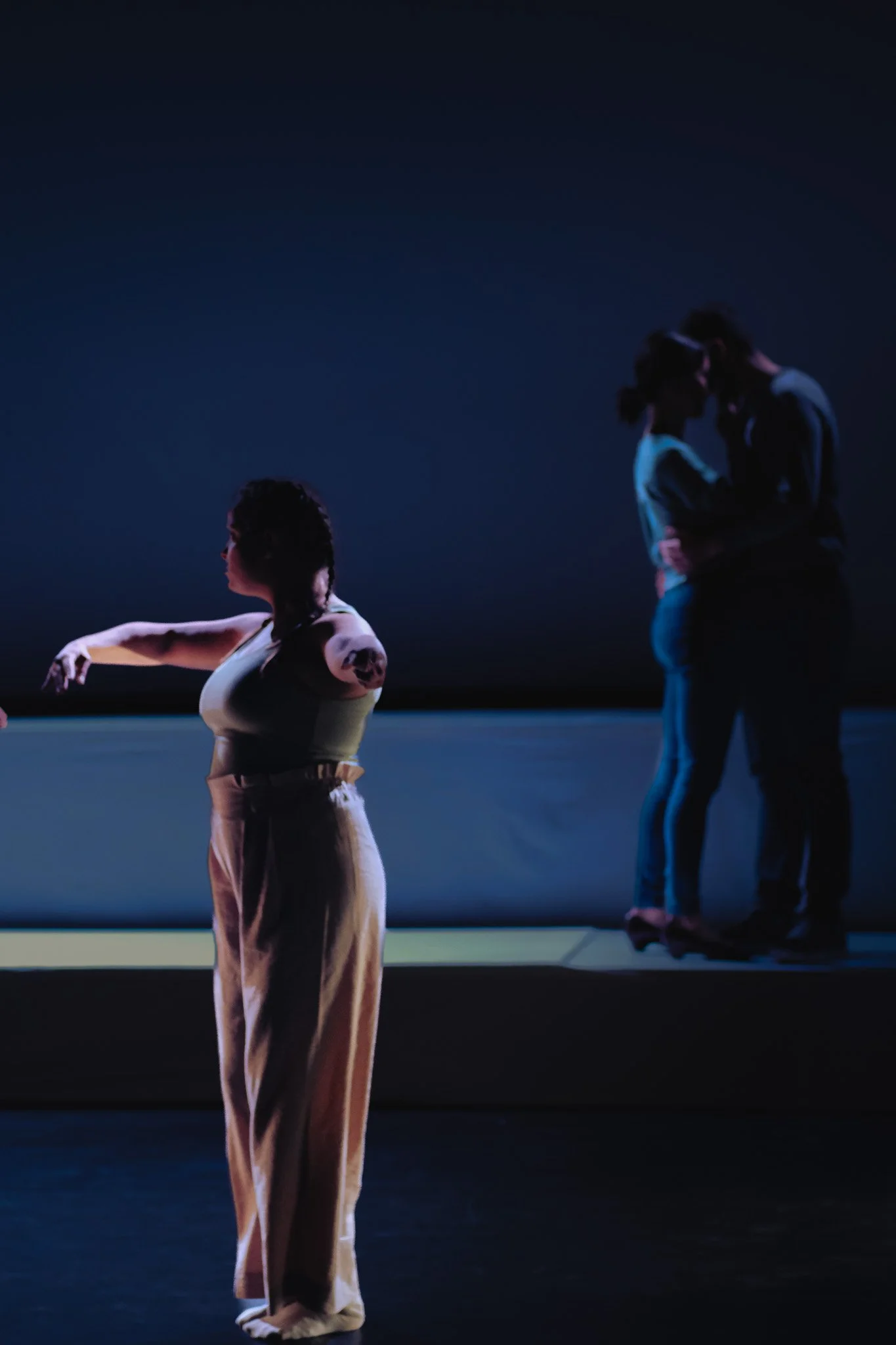 Drei Menschen auf einer Bühne, zwei küssen sich im Hintergrund, eine Person im Vordergrund schaut weg und tanzt mit für sich, bei gedämpftem Licht.