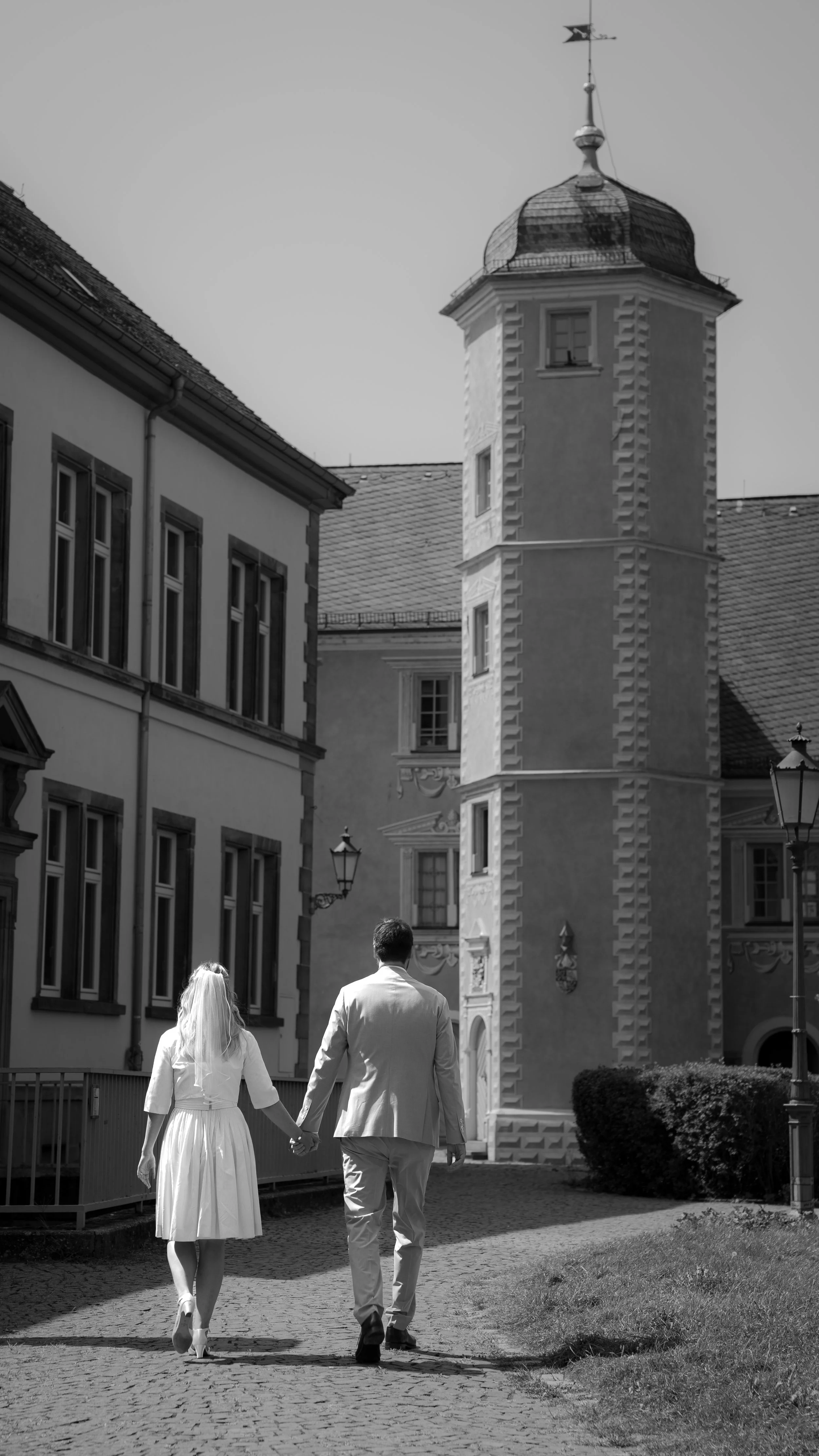 Ein Paar hält Händchen und geht auf einem gepflasterten Weg in einem historischen Stadthaus, alles in Schwarz-Weiß.