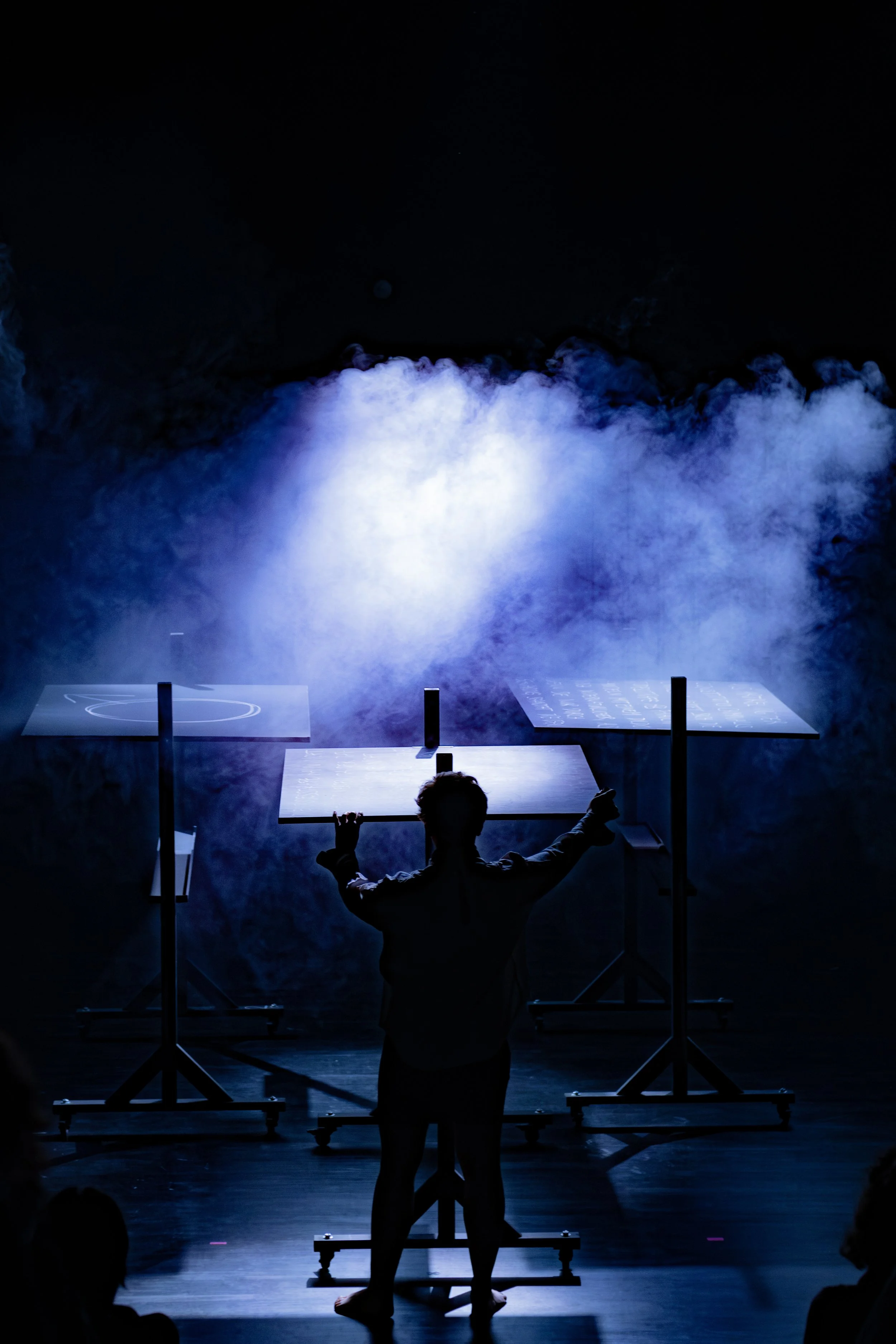 Ein Mensch steht vor drei Tischen auf einer Bühne, während blauer Rauch oder Nebel im Hintergrund aufsteigt, beleuchtet mit blauem Licht, bei einer Aufführung oder Performance.