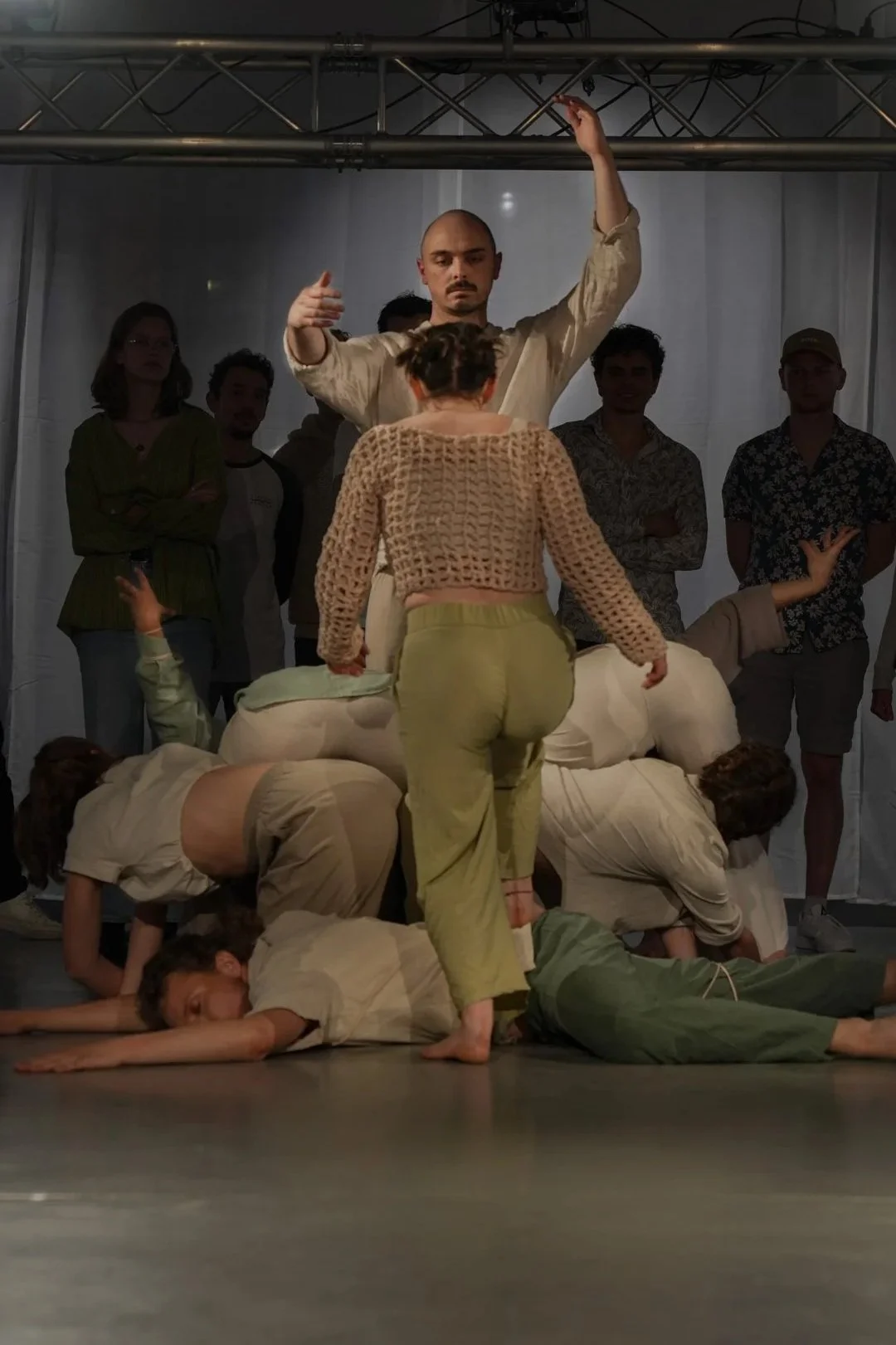 Menschen in einer Performance, die auf einem offenen Boden eine Szene mit verschiedenen Positionen und Gesten darstellen, im Hintergrund Zuschauer und eine einfache Theaterkulisse.