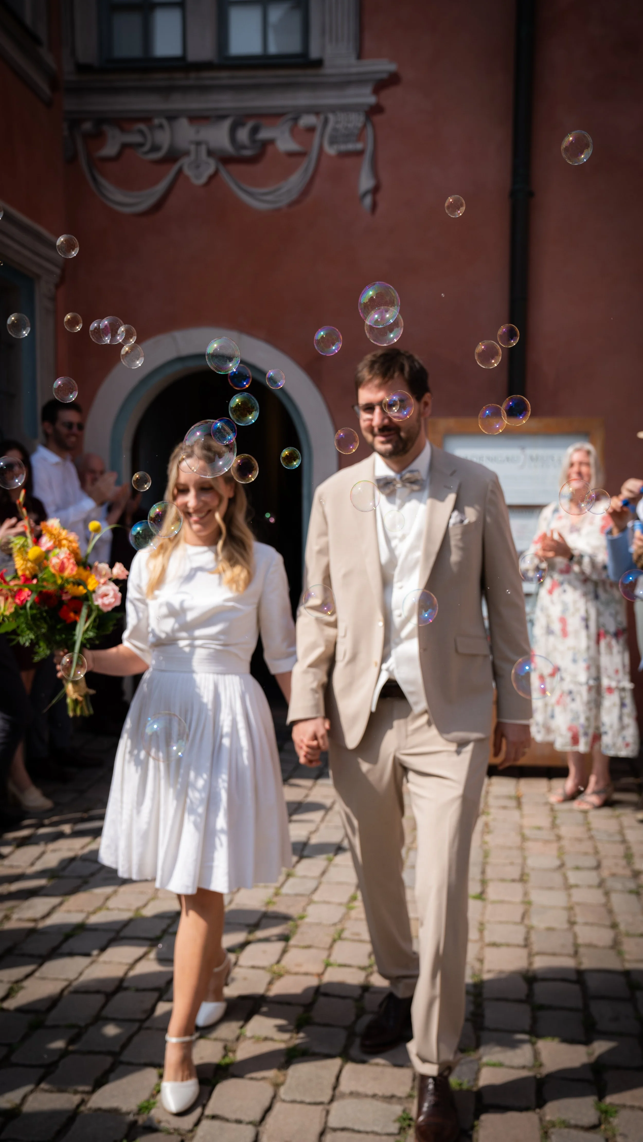 Ein brautpaar läuft Hand in Hand auf Kopfsteinpflaster vor einer Gruppe von Hochzeitsgästen, während Seifenblasen in der Luft schweben, bei einem festlichen Anlass im Freien, vor einer historischen roten Gebäudefront mit dekorativen Elementen.