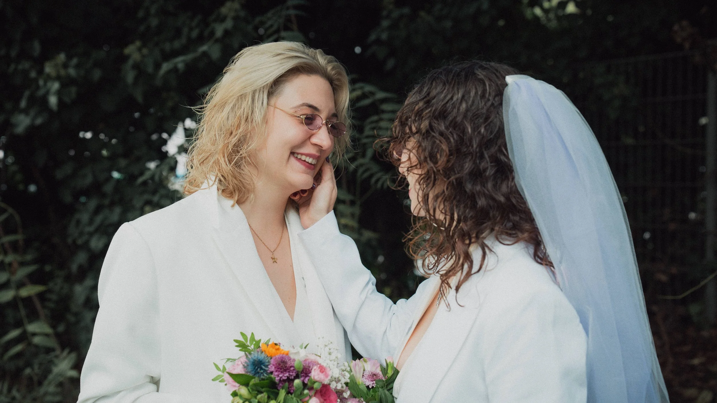 Zwei Frauen bei einer Hochzeit im Freien, eine trägt einen weißen Anzug und hält einen Blumenstrauß, die andere trägt einen weißen Brautkleid mit Schleier.
