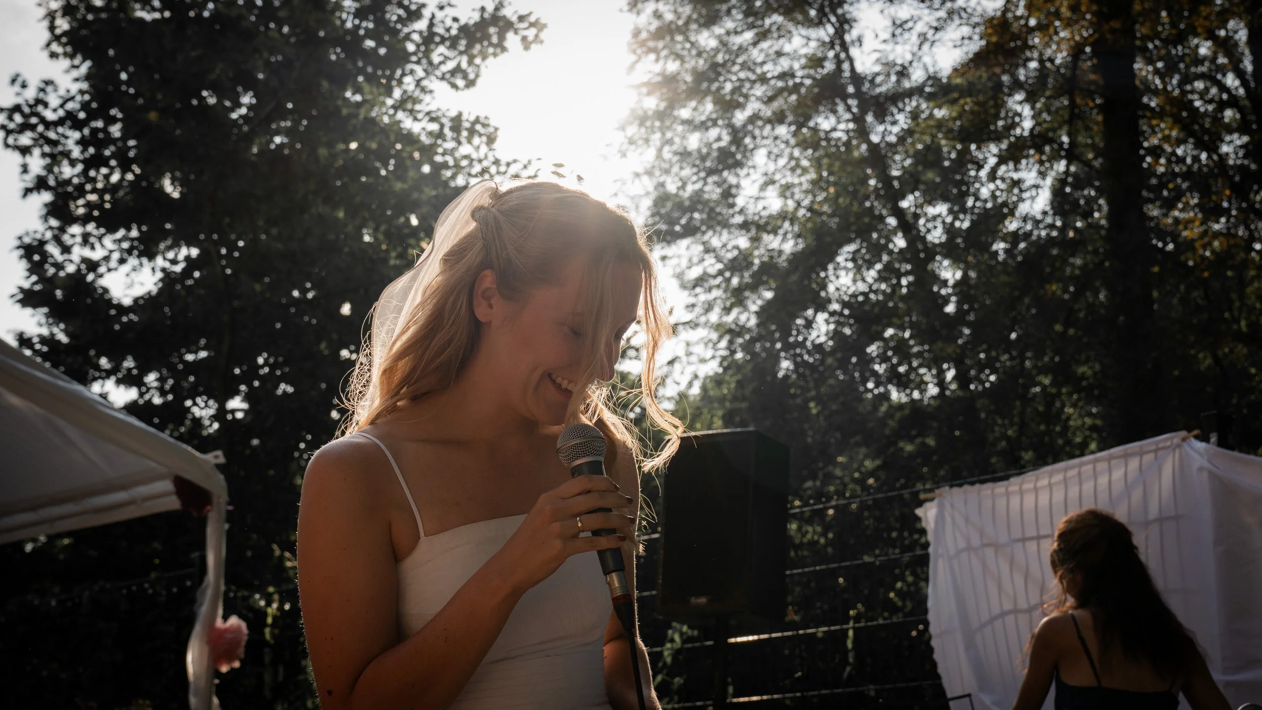 Eine lachende Frau in weißem Kleid hält ein Mikrofon bei einer Outdoor-Hochzeit im Sonnenlicht, umgeben von Bäumen.