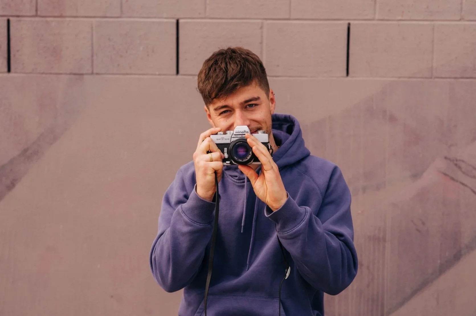 Junger Mann mit braunen Haaren in einem blauen Hoodie, der eine Kamera hält und in die Linse blickt, vor einer grauen Wand.