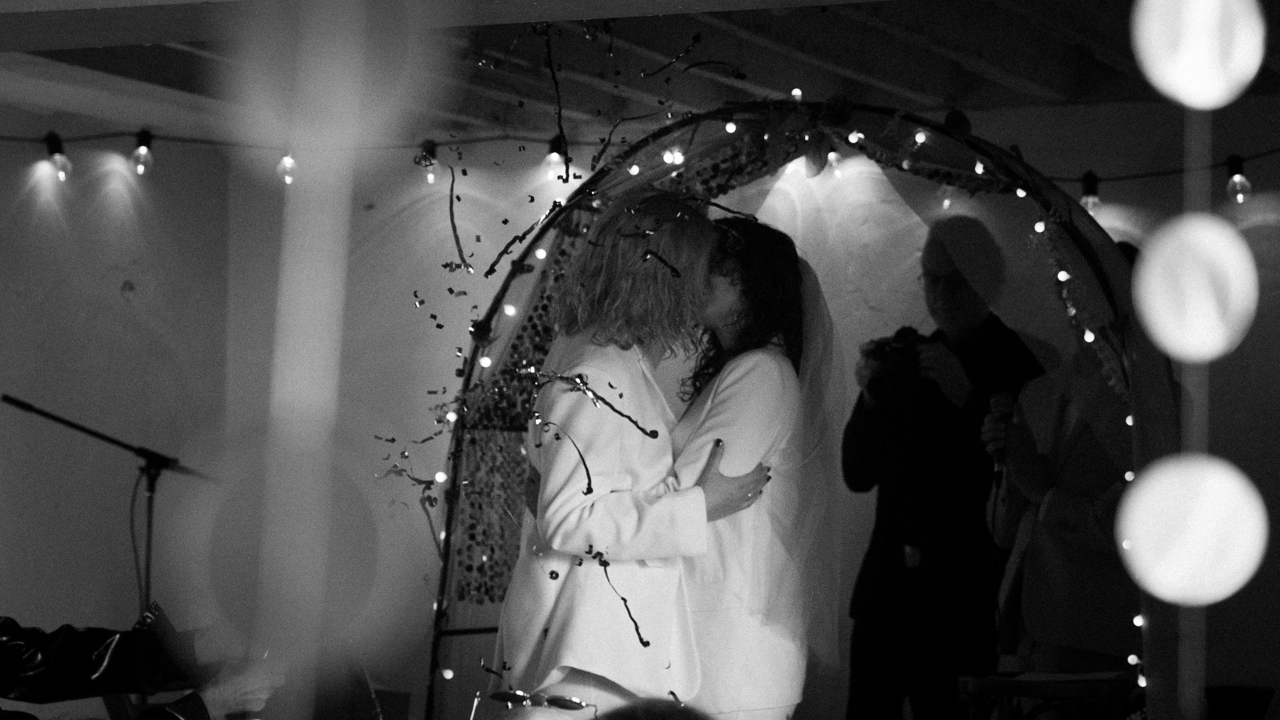 Zwei Frauen küssen sich unter einem Traubogen bei einer Feier, im Hintergrund stehen zwei Männer in einem dunklen Raum.