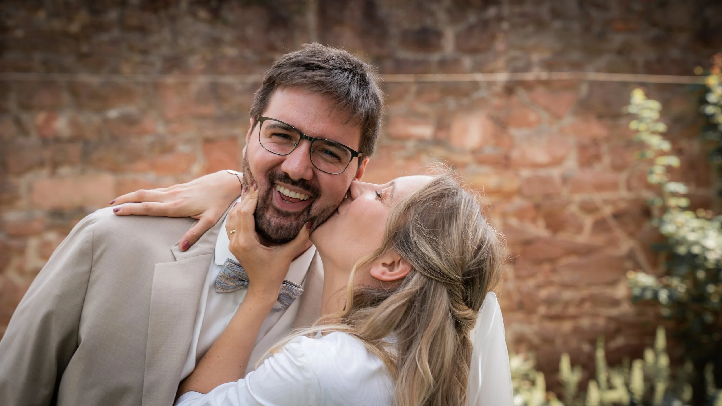Ein lachendes Paar bei einer Hochzeit, der Mann trägt eine Anzugjacke und eine Fliege, die Frau küsst ihn auf die Wange, vor einer Backsteinmauer im Freien.