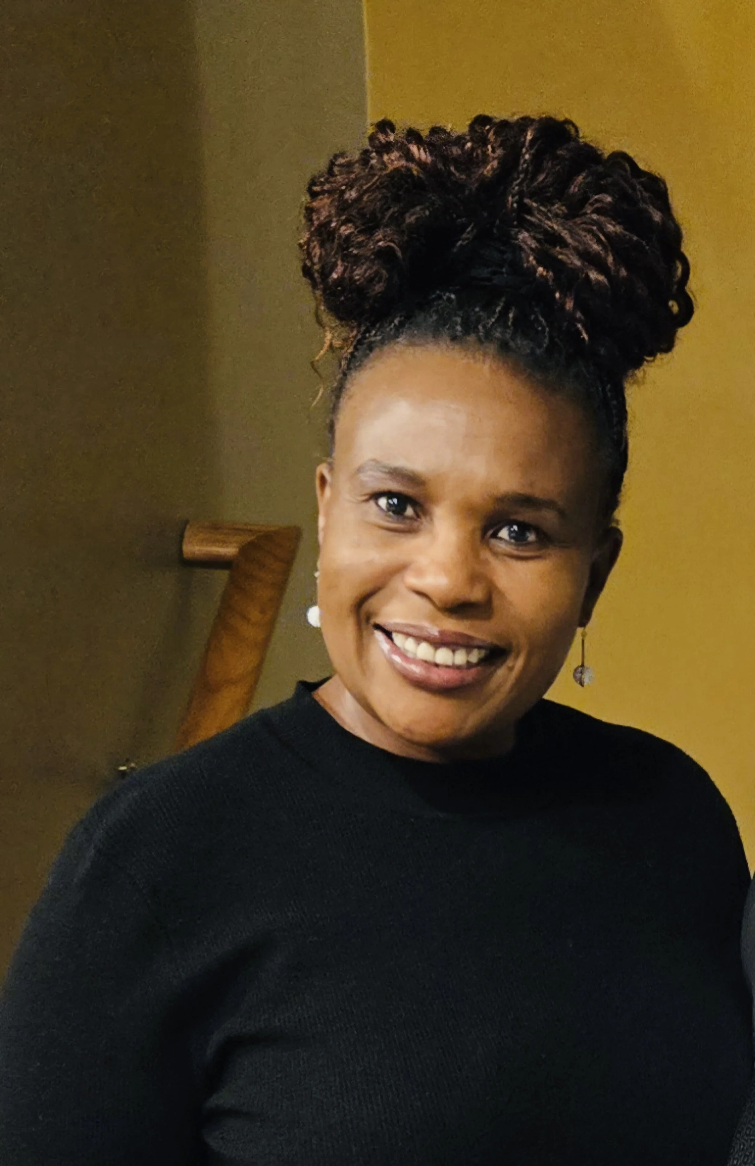 A woman with dark skin, styled hair in large curls, wearing a black top and earrings, smiling in front of a yellow wall.