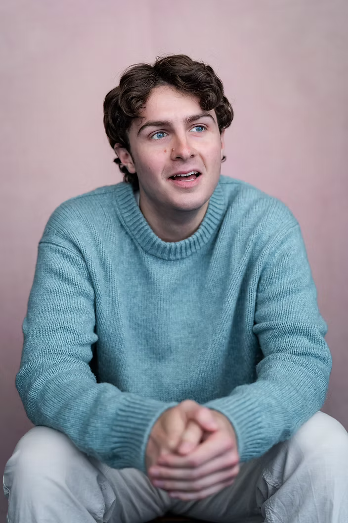 A young man with curly brown hair and blue eyes, wearing a light blue knit sweater, sitting with hands clasped in front of him against a pink background, looking slightly off-camera with a thoughtful expression.