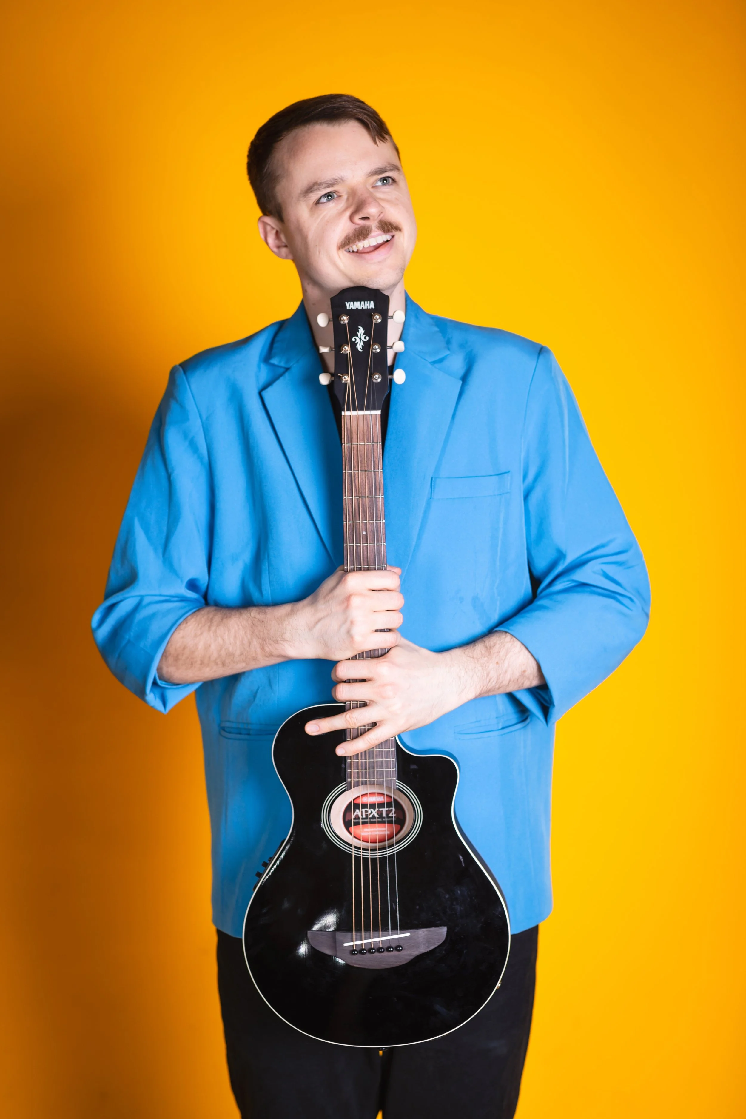 A smiling man in a bright blue blazer holding a black acoustic guitar against a yellow background.
