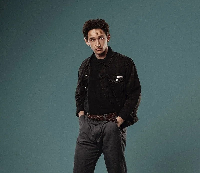 A man with curly hair wearing a black denim jacket, black shirt, and dark gray pants stands against a plain backdrop, with hands in pockets and a skeptical expression.