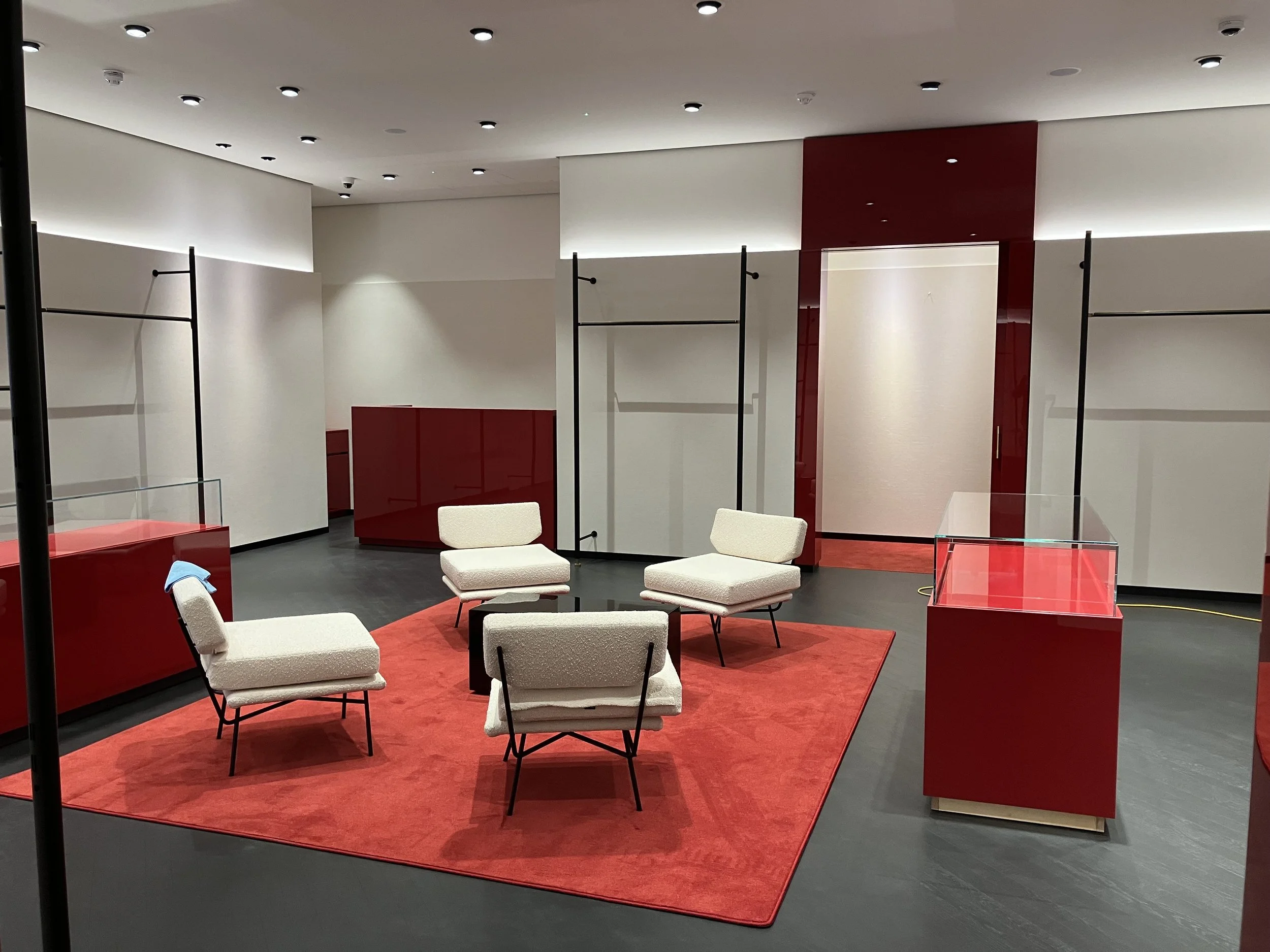 A modern interior space with white walls, a red area, and a seating arrangement on a red rug. The room features ceiling lights and some black metal and glass partitions.