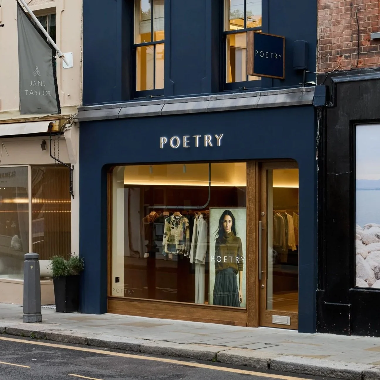 Storefront for a clothing store named 'Poetry,' featuring a large window display with a poster of a woman and clothing inside, situated on a city street with neighboring buildings and sidewalk.