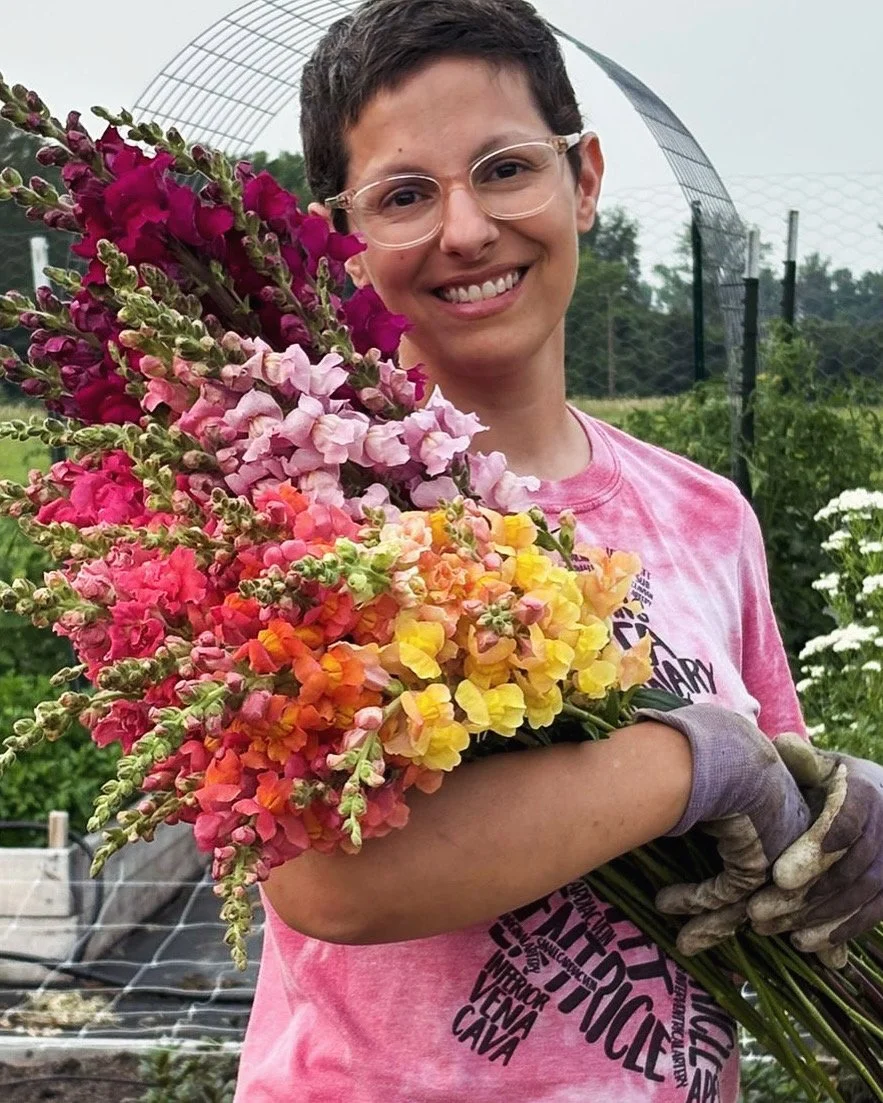 A smiling person with short hair, glasses, and a pink tie-dye shirt is holding a large bouquet of colorful snapdragon flowers in a garden.