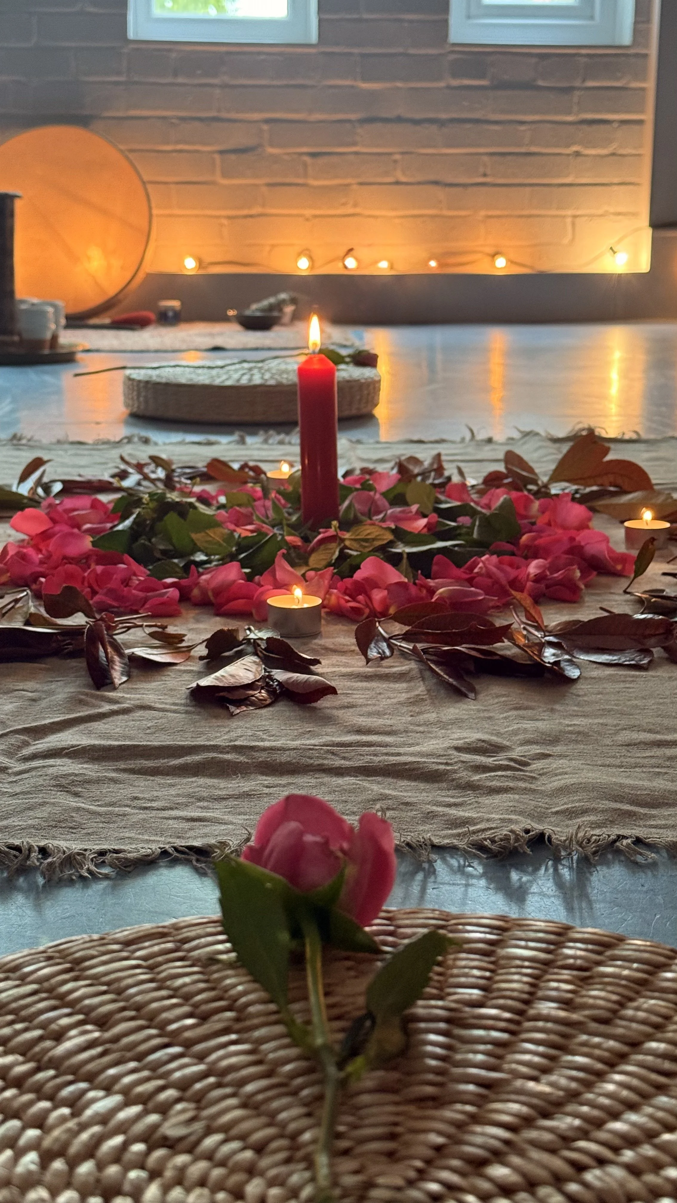 A cozy indoor setting with a candlelit centerpiece on a table. The centerpiece features a pink candle surrounded by rose petals and leaves. In the foreground, a pink rosebud is placed on a woven mat. The background includes a brick wall, small windows, and warm string lights, creating an inviting ambiance.