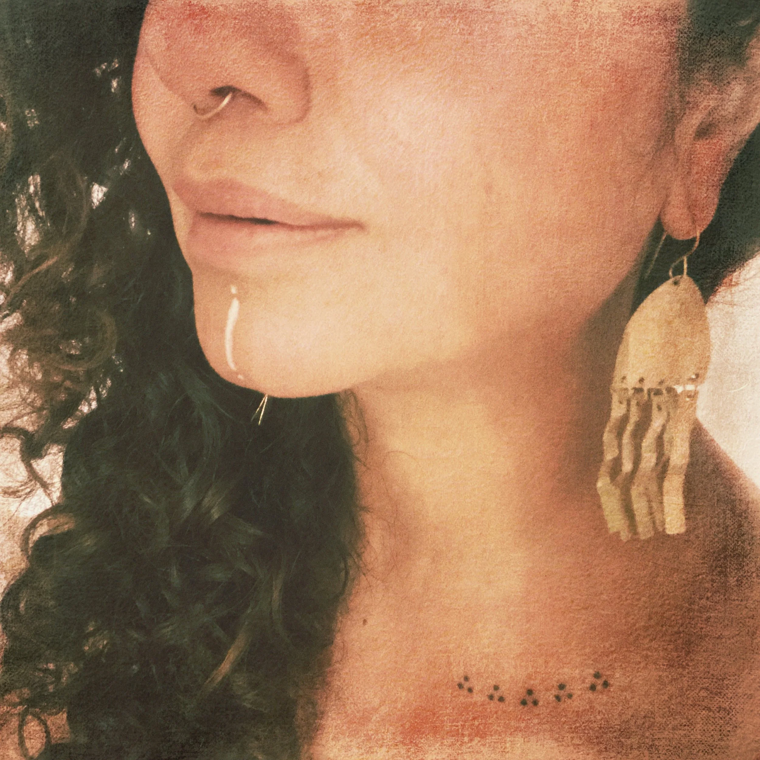 Close-up of a woman's face showing her nose, lips, and part of her curly hair, with a tattoo of small black dots on her collarbone and an ornate earring hanging from her left ear.