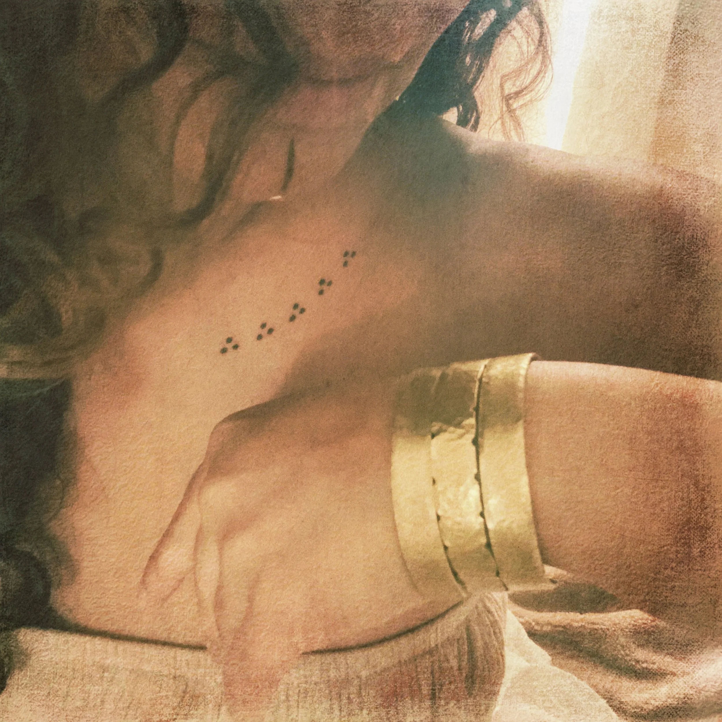 A woman with long hair topless, wearing gold jewelry on her wrist, showing a tattoo of small dots and flowers on her side.