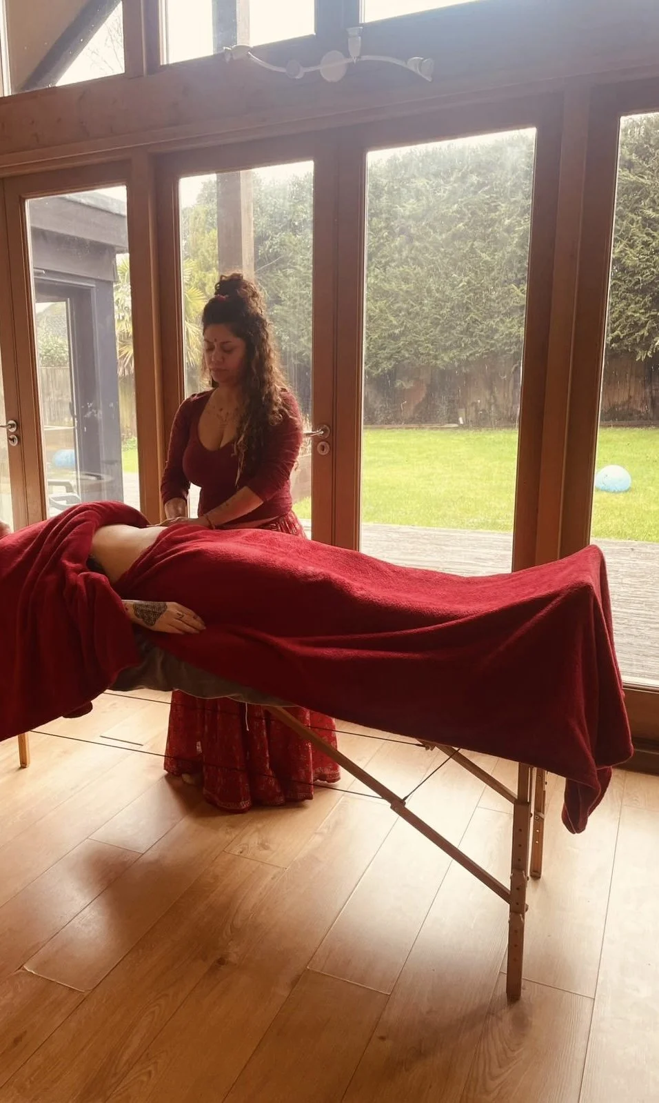 A woman giving a massage on a massage table covered with a red blanket in a room with large windows and a view of a grassy backyard.