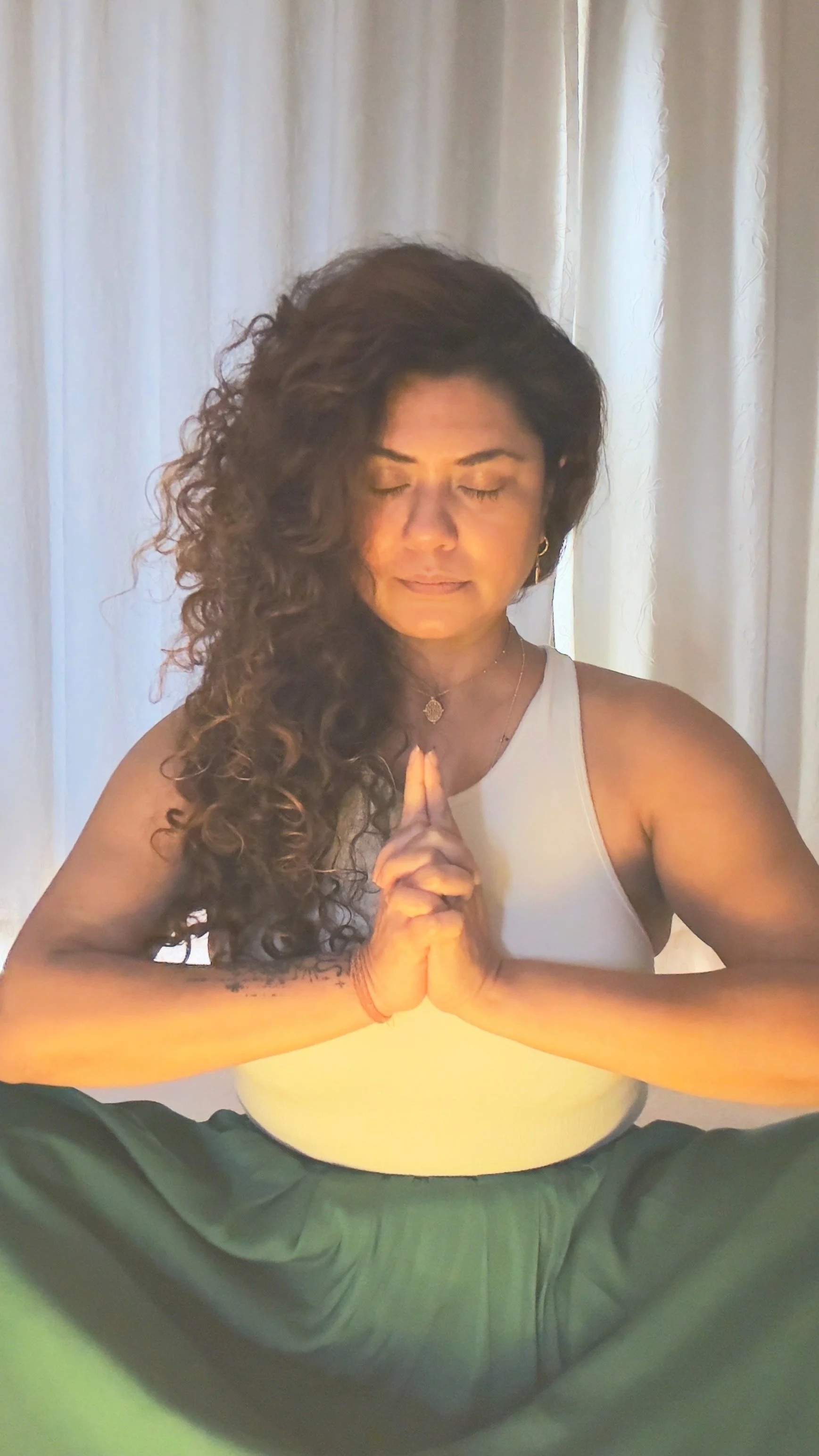 A woman with long, curly hair sitting cross-legged in a yoga pose with hands pressed together, eyes closed, in front of a white curtain.