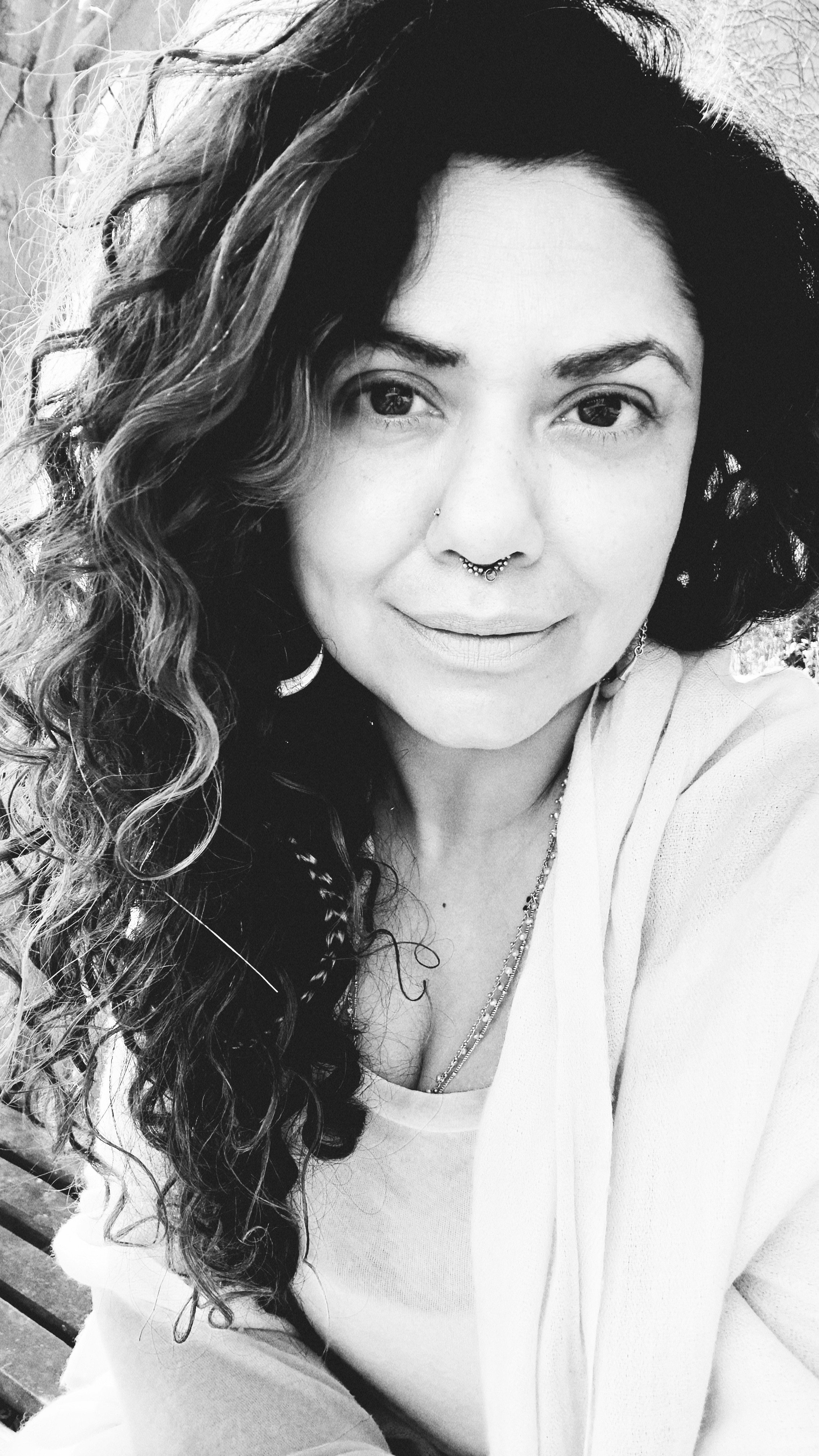 Black and white close-up of smiling woman with curly hair, septum piercing, wearing a necklace and light-colored top, seated on a bench outdoors.