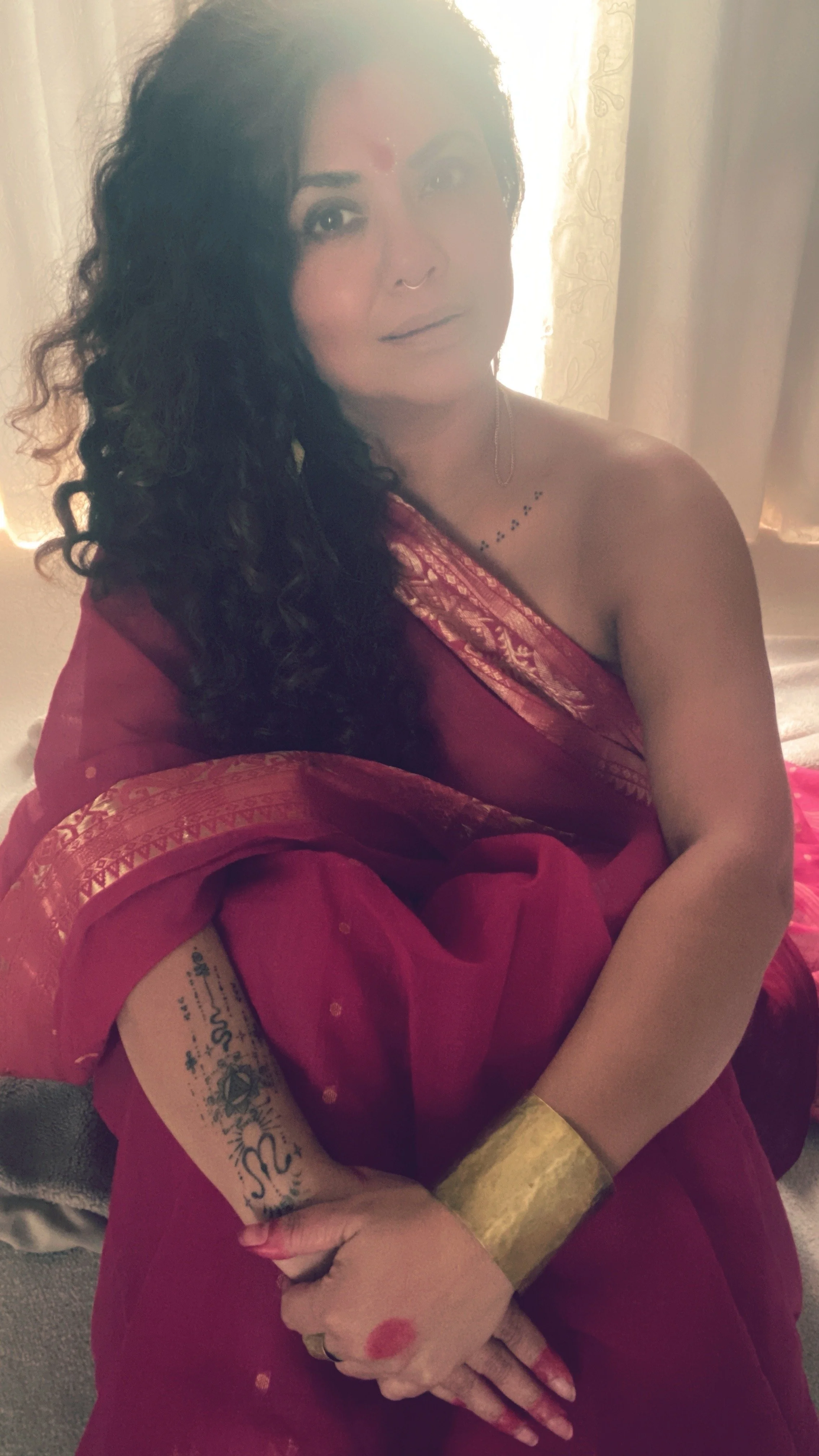 Woman with dark curly hair wearing traditional red sari with gold accents, sitting on the floor near sunlight with arms crossed and gold jewelry, including a bracelet and ring, and tattoos on her arm and shoulder.
