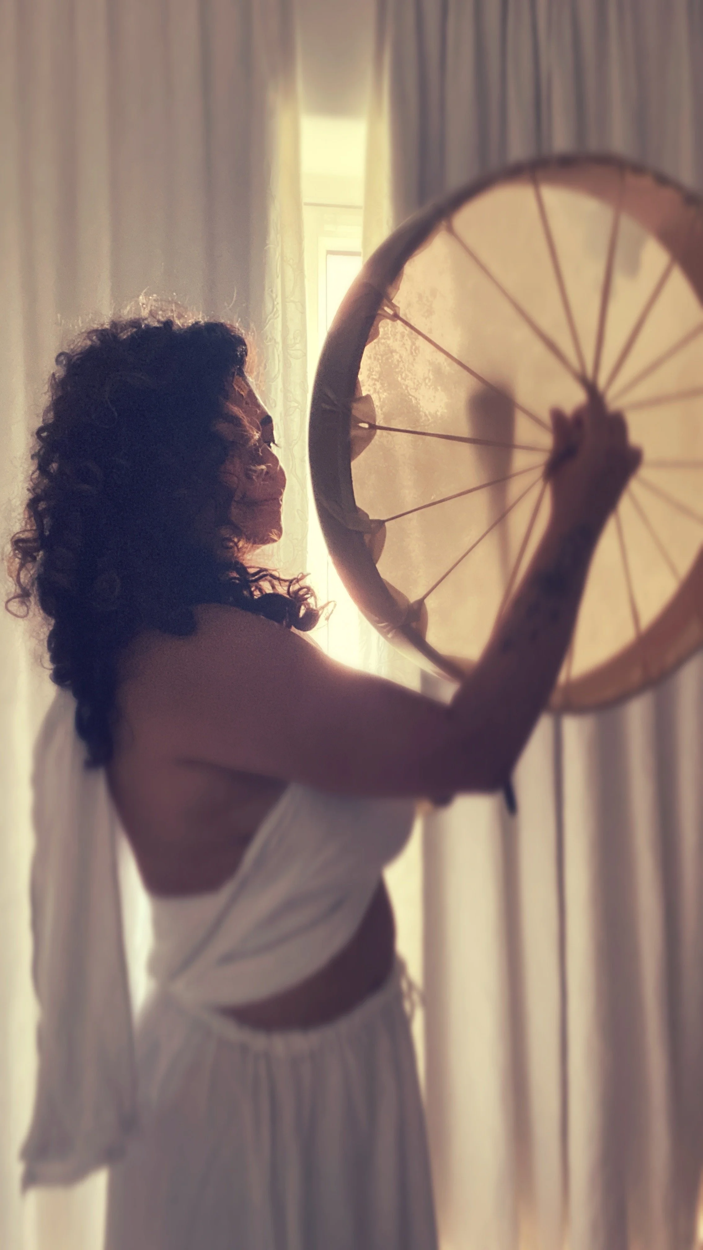 A woman with curly hair in a white dress holding a large, closed umbrella in front of a window with curtains, illuminated by sunlight.