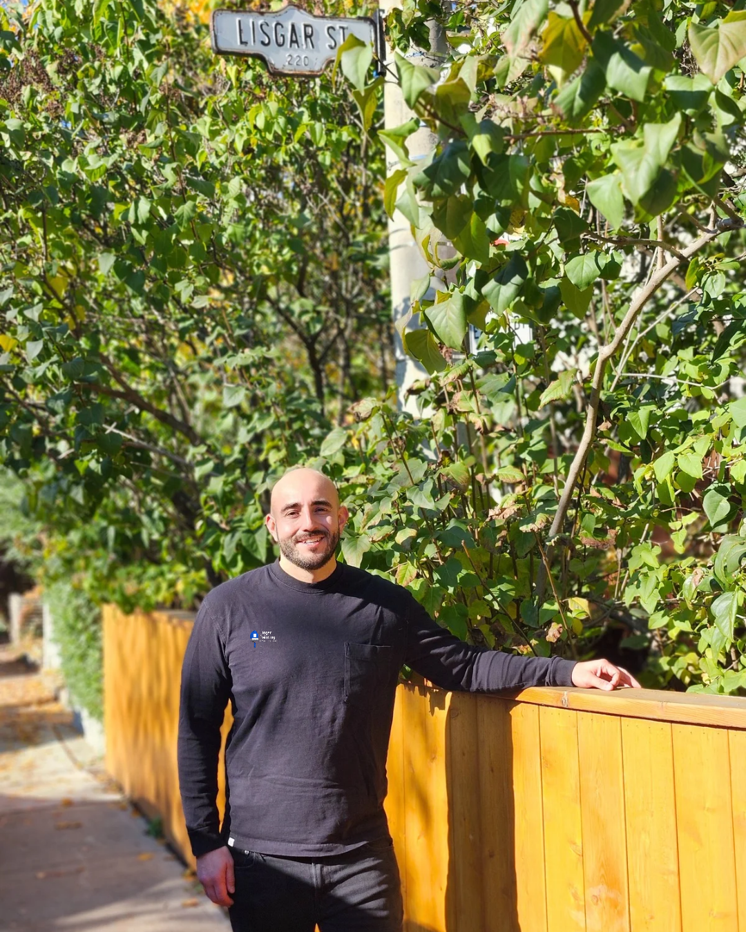 Man standing outdoors near a wooden fence, with lush green leafy bushes in the background and a street sign that reads 'LISGAR ST'.