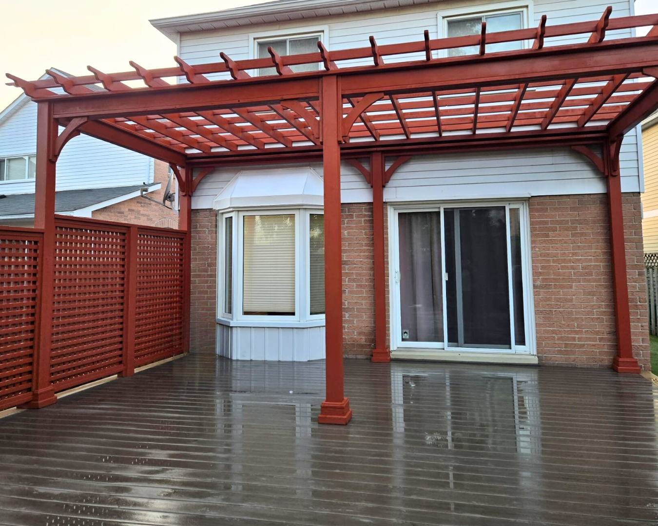 A wooden deck attached to a house with a white upper level and brick lower level, featuring a red lattice privacy screen on the left side and a red pergola structure above.