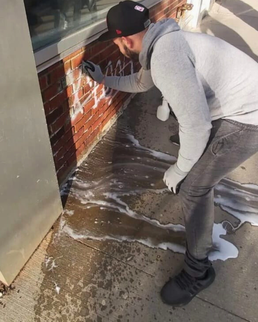 A man wearing a gray hoodie, gray jeans, black shoes, a black cap, and gray gloves, is cleaning graffiti off a brick wall with a spray bottle on the ground and soap suds on the concrete sidewalk.